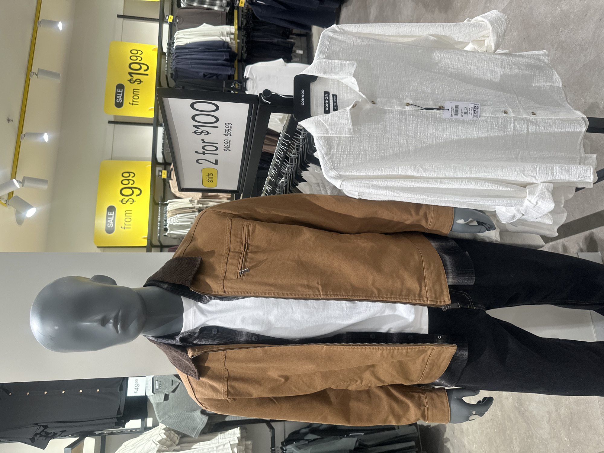 The image depicts a retail store display featuring mannequins and clothing. A male mannequin on the left is dressed in a brown jacket layered over a plaid shirt and a white t-shirt, with dark jeans. To its right, another mannequin is displayed wearing a white long-sleeved shirt. Behind the mannequins, racks of clothing are visible, and prominent yellow sale signs hang on the wall. One sign advertises "SALE from $9.99," and another "SALE from $19.99." A black and white sign with text "shirts 2 for $100" is also present. The store environment appears to be a clothing department within a larger retail space. The location is identified as Coffs Harbour, Australia.