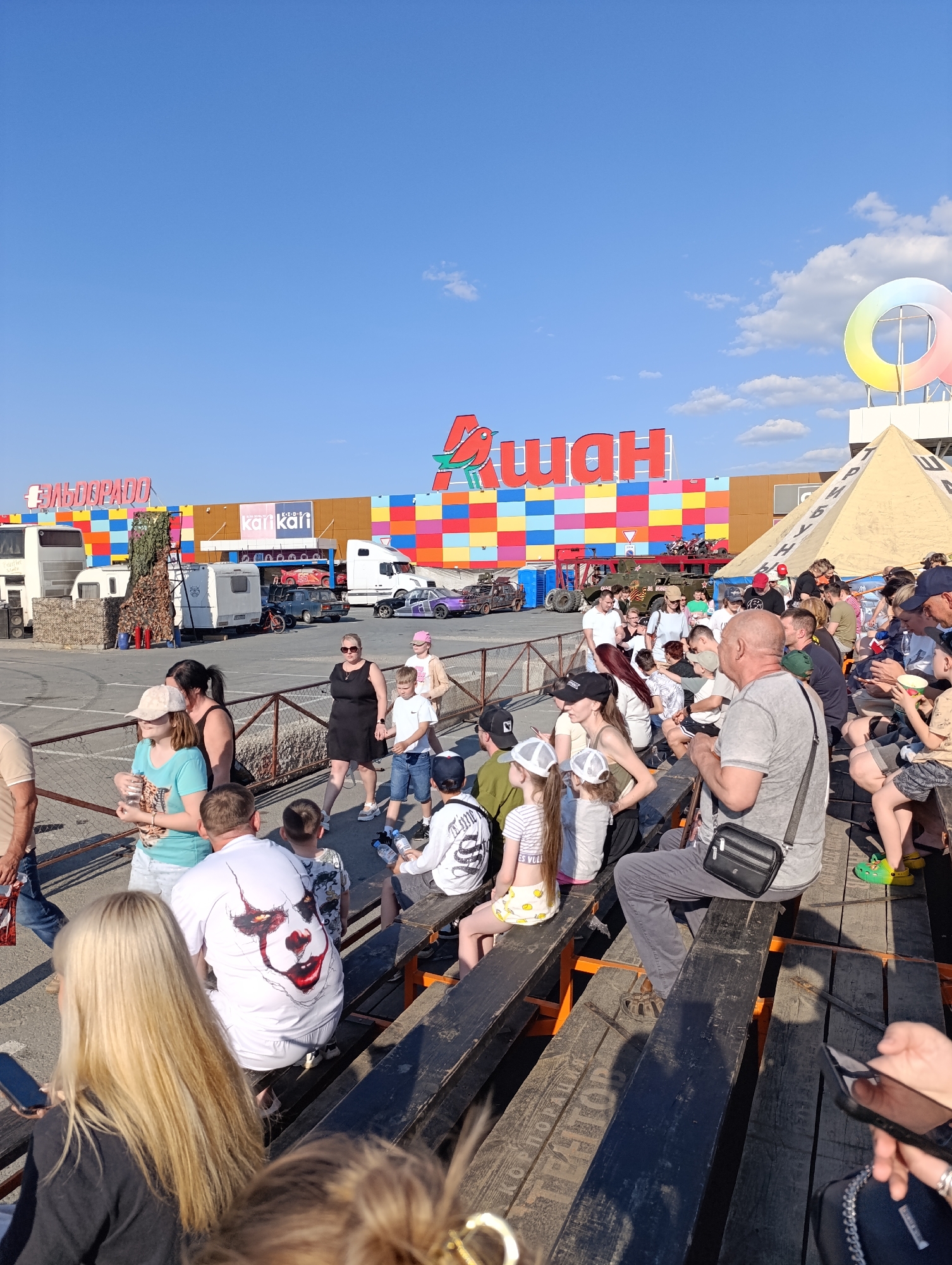 The image depicts a crowd of people gathered in an outdoor setting in Unknown, Unknown. They are seated on wooden benches, appearing to watch something beyond the frame. In the background stands a large building with a colorful facade, featuring the word "АШАН" (Auchan) in prominent red letters. To the left of the Auchan building is a sign for "ЭЛЬДОРАДО" (Eldorado). There are various vehicles parked in front of the building, including a white truck and several modified cars. A pyramid-shaped structure with lettering on its side is visible to the right of the main building, topped with a large, colorful circular logo. The sky above is blue with scattered clouds. The scene appears to be taking place during the daytime, with sunny weather conditions.
