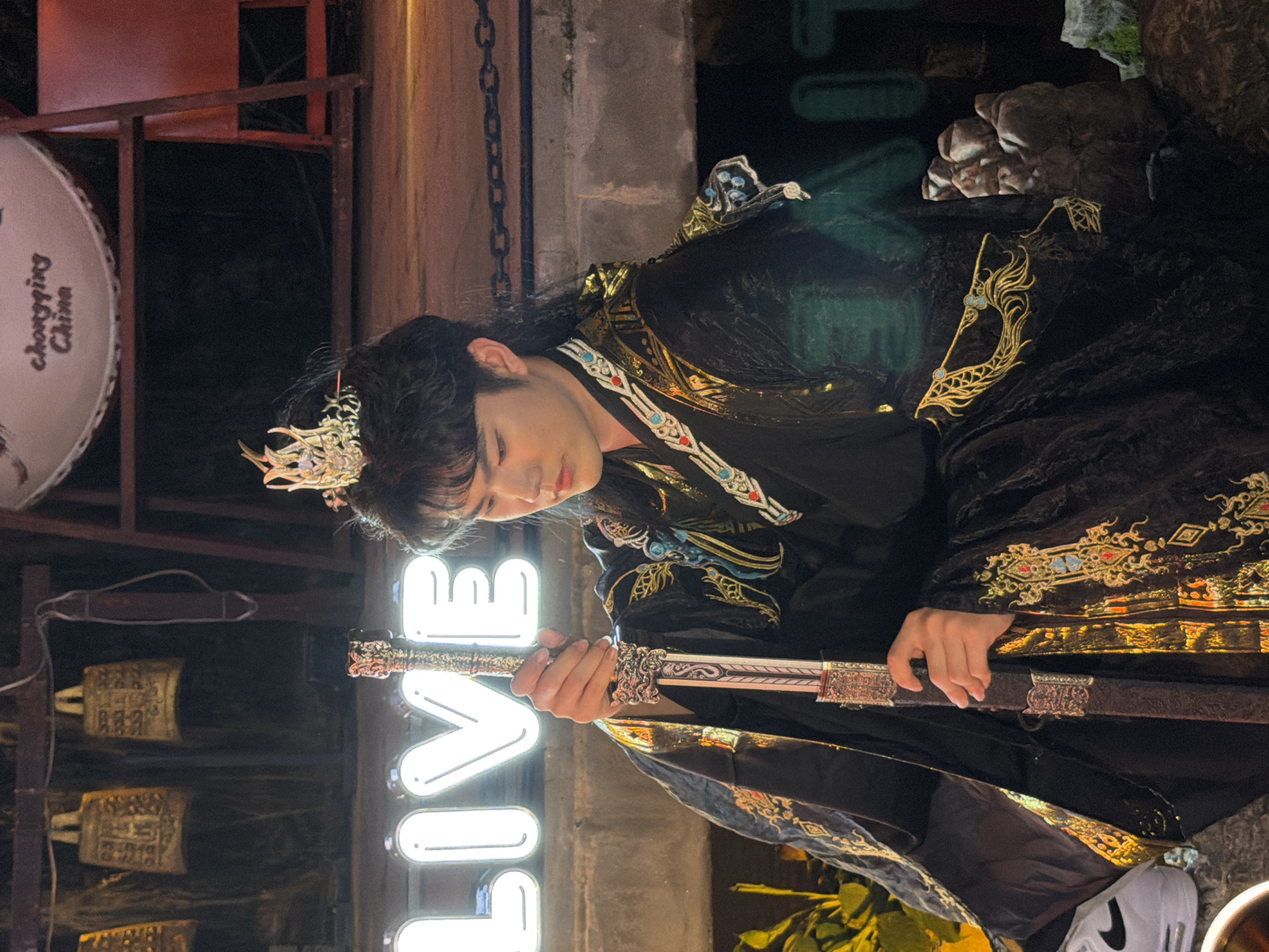 In Yuzhong District, China, a person dressed in ornate traditional Chinese attire, complete with a crown, holds a decorative sword. The individual has dark hair and is looking downwards. The background features neon signs, one of which spells out "LIVE" in large white letters. A large drum with the text "Chongqing China" is also visible. Several golden bells hang in the upper left portion of the frame. The setting appears to be outdoors at night, possibly a street or performance area, with various textures and objects suggesting a dynamic urban environment.