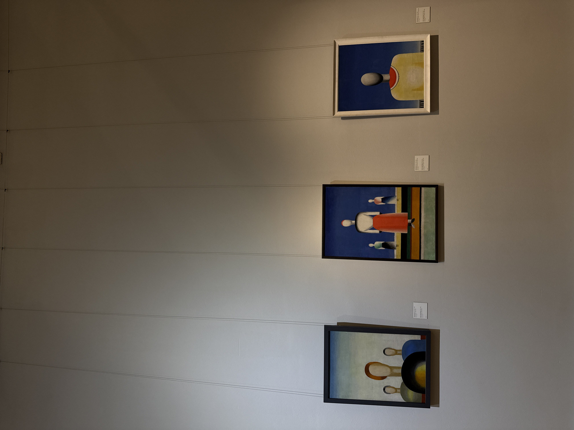 Three paintings are hung on a light gray wall, one above the other. Each painting has a small white label above it. Thin wires suspend the paintings. The top painting has a white frame and features a blue background with abstract shapes, including a white ball and yellow and red lines. The middle painting has a dark frame and depicts abstract figures in red, white, green, and black against a blue background. The bottom painting has a dark frame and shows abstract shapes in blue, black, yellow, and brown against a light background. The location is likely an art gallery or museum in Moscow, Russia, but the specific details that indicate this are not present in the image.