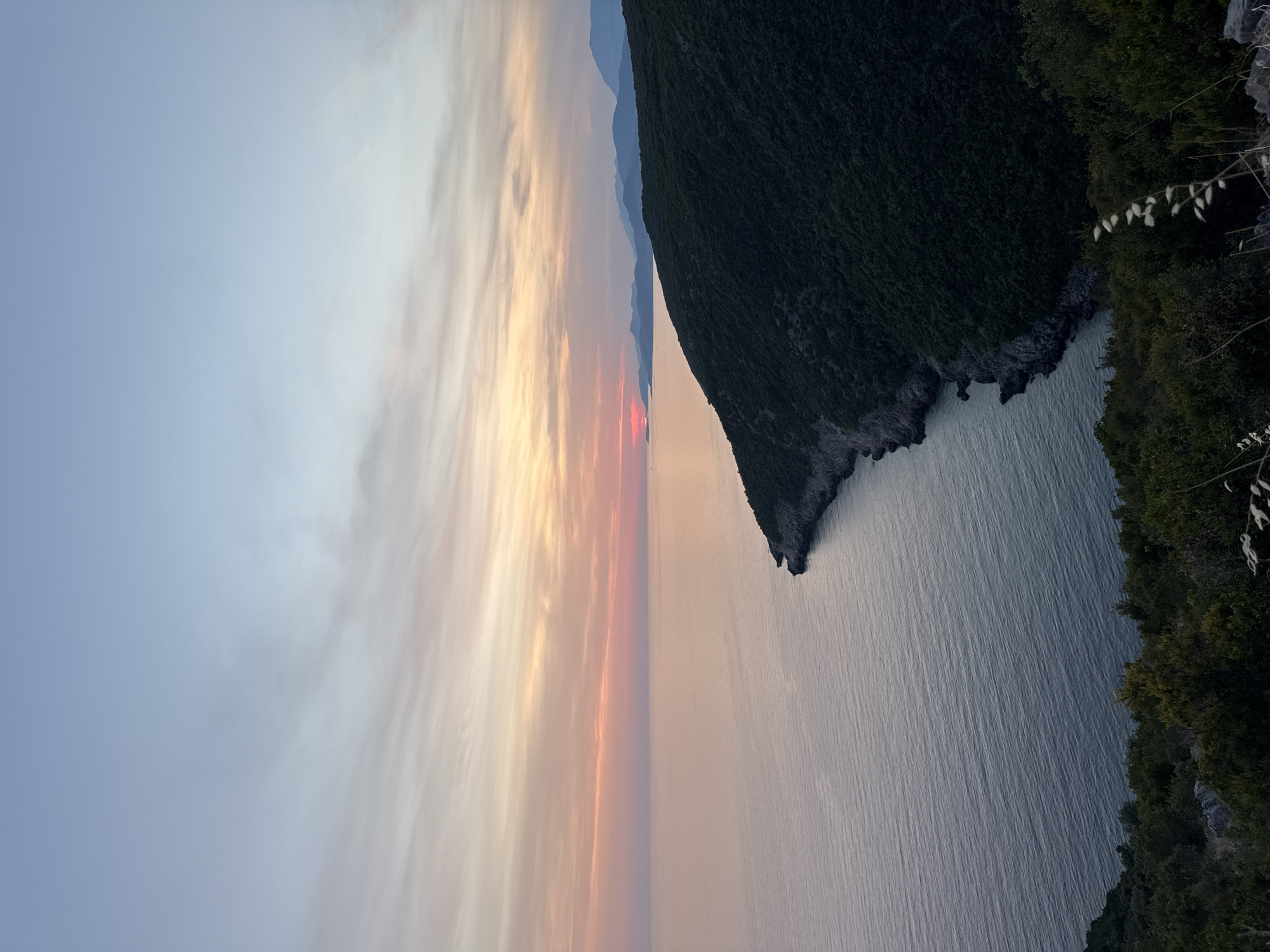 The image depicts a sunset over the Adriatic Sea, with a forested coastline in the foreground and distant mountains. The sky is a gradient of soft blues, pinks, and oranges, with wispy clouds. The sea is calm and reflects the colors of the sky. The coastline is covered in dense green vegetation, and the rugged terrain is visible along the water's edge. The scene is devoid of people or man-made structures, conveying a sense of natural beauty and tranquility. The location is identified as the city of Zagora, Montenegro, a coastal region known for its scenic landscapes.