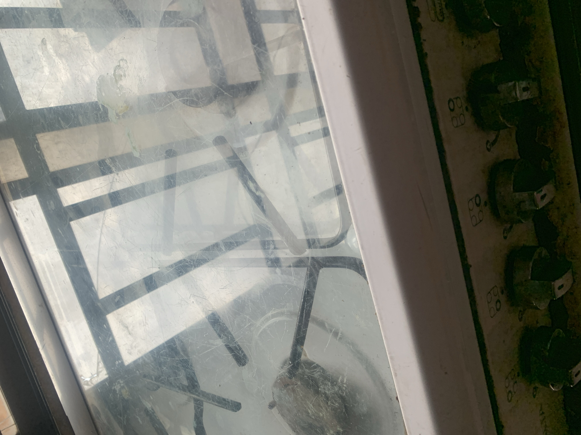 The image shows a close-up, interior view of a dirty gas stove. The surface is covered in scratches and there are food stains and grease. The grate of the stove is visible, along with one of the burners. The control knobs of the stove are also visible at the bottom of the image and appear to be covered in grease and grime. There are symbols above each knob indicating the burner they control. The overall impression is of a well-used and neglected appliance. This image is likely located in Jalingo, Nigeria, given the prompt.