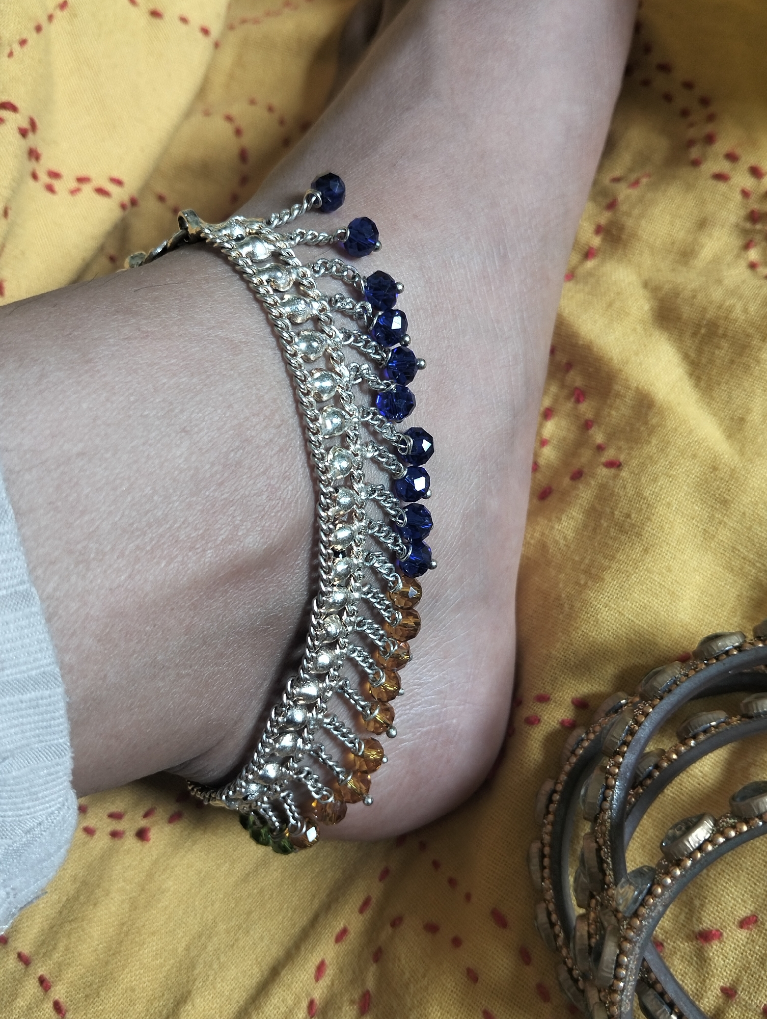 An ankle with jewelry is shown against a yellow textured fabric. The ankle is adorned with a silver-colored anklet featuring dangling beads in varying colors, including dark blue, amber, and green. Additional silver-colored jewelry is located next to the ankle. The environment is a yellow fabric with stitch-like patterns. The image was taken indoors. The location is unknown, no information suggests the city of Dhaka, Bangladesh.