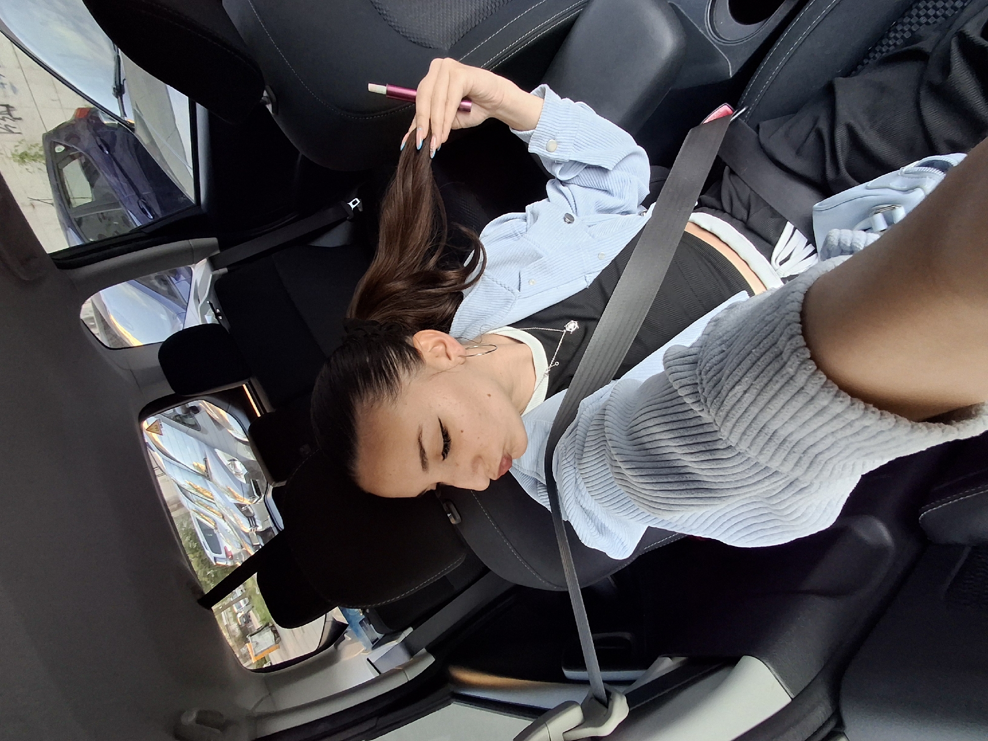 The image shows a young woman lying horizontally across the passenger seat of a car, viewed from an overhead perspective. Her head rests on the headrest, and she is secured by a seatbelt. She has dark hair styled in a ponytail and wears a light blue, ribbed long-sleeved shirt over a dark top with white and orange stripes. Her fingernails are painted light blue, and she holds a magenta or purple pen-like object (possibly a vape pen or stylus) in her right hand, near her hair. Her eyes appear closed, suggesting she is resting or posing. Freckles are visible on her face.
The car's interior features dark grey and black seats and visible seatbelts. A light grey ribbed sleeve, matching the texture of her blue shirt, is prominent in the foreground. A light blue bag is partially visible near her legs.
Reflections in the car windows and mirrors reveal a sunny daytime outdoor setting, indicating clear weather. These reflections show parts of other vehicles (including a dark blue car with an open door and a white car), some greenery, and buildings. What appears to be graffiti or markings on a wall is also faintly visible in one reflection. A small triangular symbol is also reflected. The scene depicts a casual moment inside a car, likely in an urban or suburban environment, consistent with Sofia, Bulgaria. No legible text is visible within the frame.