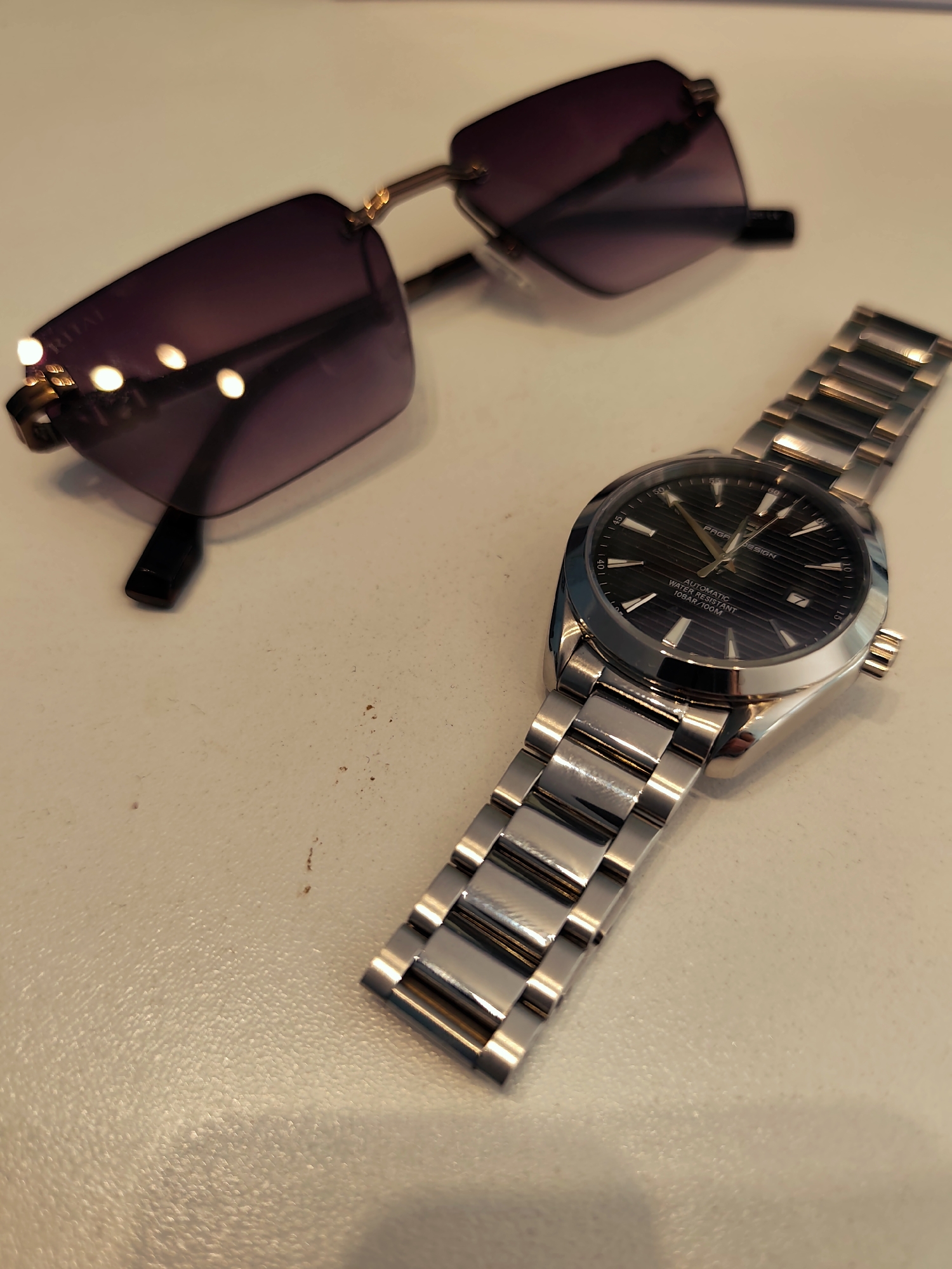 A pair of sunglasses and a wristwatch are arranged diagonally on a light-colored, possibly off-white, flat surface. The sunglasses are positioned in the upper left quadrant of the frame, out of sharp focus. They feature large, rimless, dark purple tinted lenses with gold-colored accents at the temples and black plastic earpieces. Reflections on the left lens show partially legible text, appearing to be "RITAT". The wristwatch is in the lower right quadrant of the frame, in sharp focus. It has a silver-toned metal case and a three-link bracelet. The watch features a black dial with a horizontal textured pattern, luminous hour markers, and hands. A date window is visible at the 3 o'clock position. Text on the dial includes "PAGANI DESIGN", "AUTOMATIC", "WATER RESISTANT", and "10BAR/100M". The surface shows minor imperfections and specks in the lower center and right, suggesting dust or texture. The scene is lit, creating reflections on the metallic and glass surfaces of both objects. No people are present.