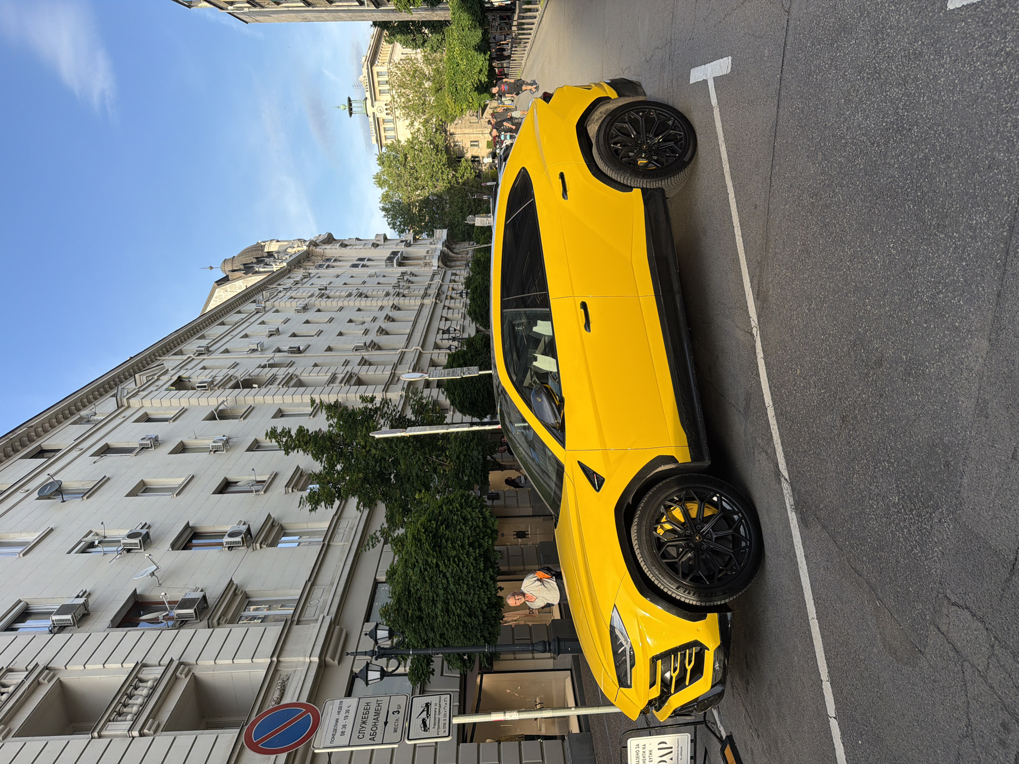 The image depicts a bright yellow Lamborghini Urus parked on a street in Sofia, Bulgaria. The car is parked along a row of white-painted buildings with multiple stories, many windows, and several visible air conditioning units. The street is paved with asphalt and features white parking lines. A parking sign is visible near the front of the car. The street also has streetlights. A few pedestrians are visible in the background, walking on the sidewalk. The sky is blue with few clouds.