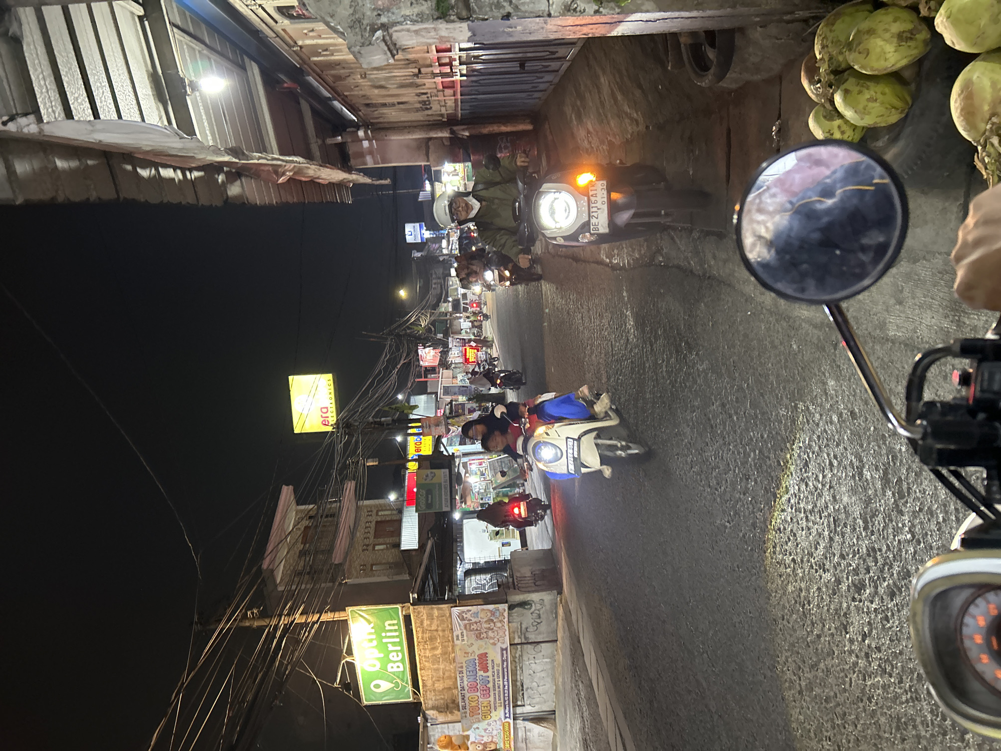 This is a nighttime, first-person perspective photograph of a street scene in Depok, Indonesia. The image is taken from a motorcycle, with the handlebar and part of the dashboard visible in the foreground. The street is paved with asphalt and illuminated by streetlights and the headlights of vehicles. Several motorcycles are visible on the road, with riders and passengers.
On the left side of the street, there are buildings with storefronts, signs, and overhead wires. A prominent green sign reads "Optik Berlin". Another sign advertises "TOKO BONERA GUEN GEPHOT JAYA". Further down the street, a yellow sign with the word "era" is visible.
On the right side of the street, there are more buildings with awnings and what appears to be a wall or fence. A pile of coconuts is visible on the ground near the edge of the street.
The overall atmosphere is that of a busy urban street at night, likely a commercial area. The lighting is mixed, with strong artificial light sources creating highlights and shadows. The image captures a slice of daily life in Depok, Indonesia.