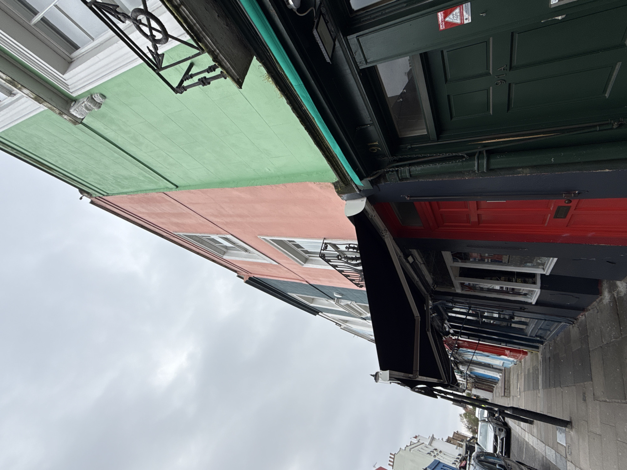 The image depicts an urban street scene on Portobello Road in Notting Hill, London, United Kingdom, under an overcast sky. The perspective is a low-angle view looking along a row of colorful terraced buildings.
On the immediate right, a multi-story building features a light green painted facade on its upper levels and a dark green ground-floor shopfront. A dark green paneled door, marked "91A" and "91", displays a "zerovision smoke barrier" sticker. An exterior light fixture is mounted above the door, beneath a narrow green awning element. White-framed windows are visible on the upper floors, with a decorative wrought-iron balcony positioned below the second-story window.
Adjacent to the green building is a multi-story structure with a pink painted facade, also featuring white-framed windows and a smaller wrought-iron balcony. Further along the row, a building with a dark blue ground floor and a red door is visible, followed by additional terraced buildings painted in various colors, including blue, red, white, and grey, receding into the distance.
A series of black retractable awnings extends over several shopfronts along the right side of the street, supported by dark poles on the paved sidewalk. The grey paved sidewalk runs parallel to the street, which contains several parked vehicles, including a silver Volvo SUV in the mid-ground. The sky above is uniformly overcast and light grey. No individuals are present within the scene.