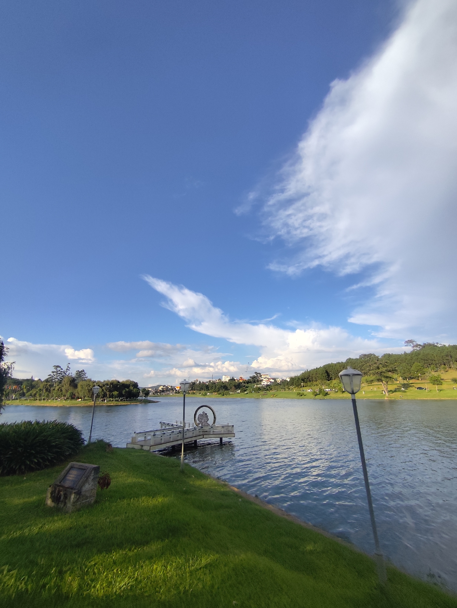 The image depicts Xuân Hương Lake in Đà Lạt, Vietnam, under a partly cloudy blue sky. The foreground features a vibrant green grassy area with a weathered stone plaque. To the right, a lamppost stands tall, its light fixture casting a shadow onto the grass.
The lake occupies the majority of the frame, its surface rippling gently. Across the water, a white pier extends outwards, featuring railings and a circular decorative element at its end, possibly a sculpture or signage. The far bank is lined with lush trees and dotted with buildings, suggesting the presence of a town or city. The distant landscape rises into hills covered in dense green forest, with some lighter-colored buildings visible on the slopes. Wispy white clouds drift across the bright blue sky, with a more prominent cloud formation stretching diagonally from the top right corner of the image. The overall impression is one of a serene natural landscape integrated with urban elements.