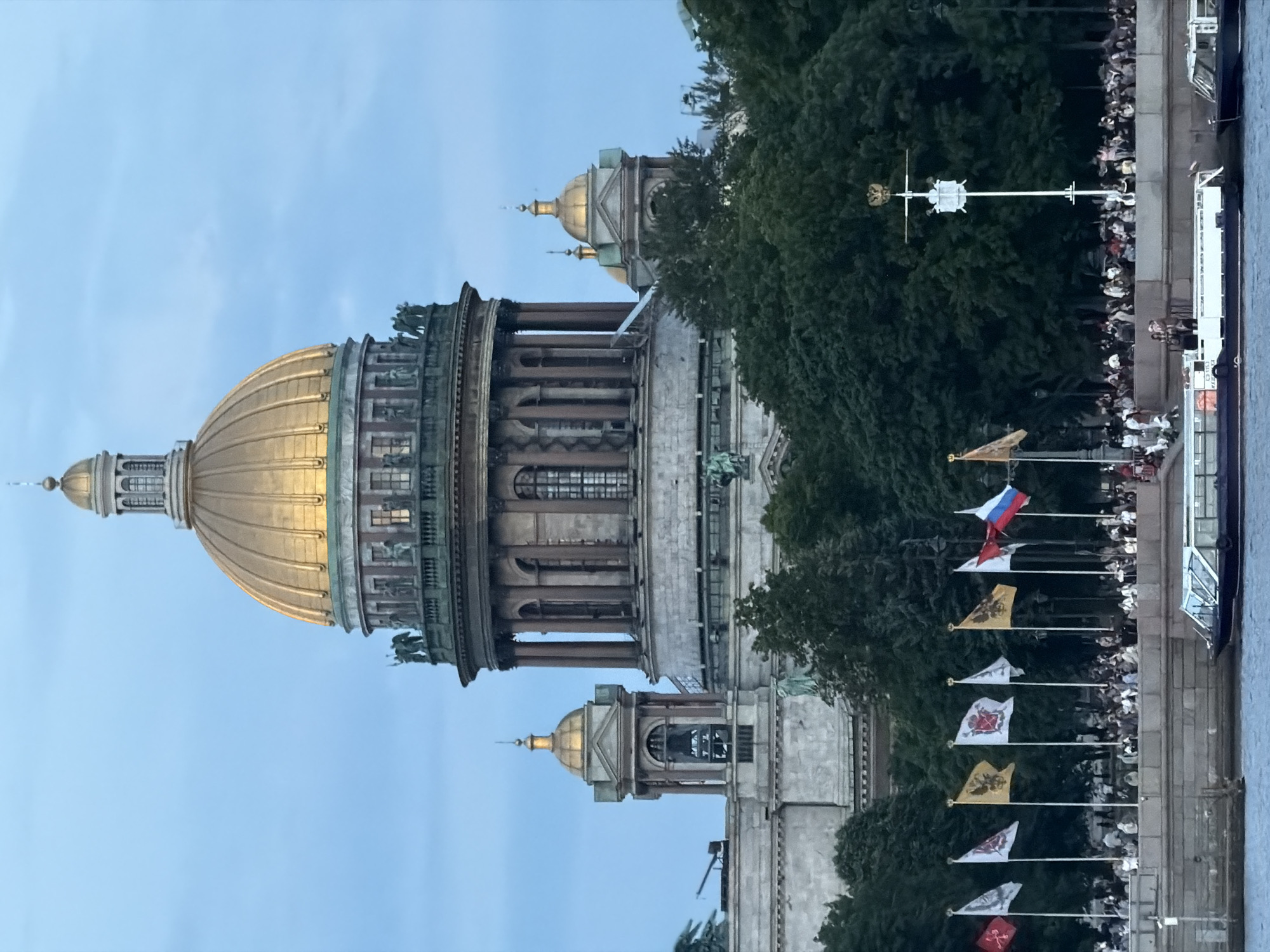 The image shows a view of St. Isaac's Cathedral in Saint Petersburg, Russia, from across a body of water. The cathedral's large golden dome is illuminated and dominates the upper center of the frame, with smaller golden domes and architectural details visible. In the foreground, a line of flags flies from poles along a waterfront promenade, where a crowd of people is gathered. Several boats, including a long, multi-decked vessel, are on the water. Dense green trees form a line behind the promenade, partially obscuring the lower levels of the cathedral. The sky is a pale blue, suggesting either early morning or late evening.