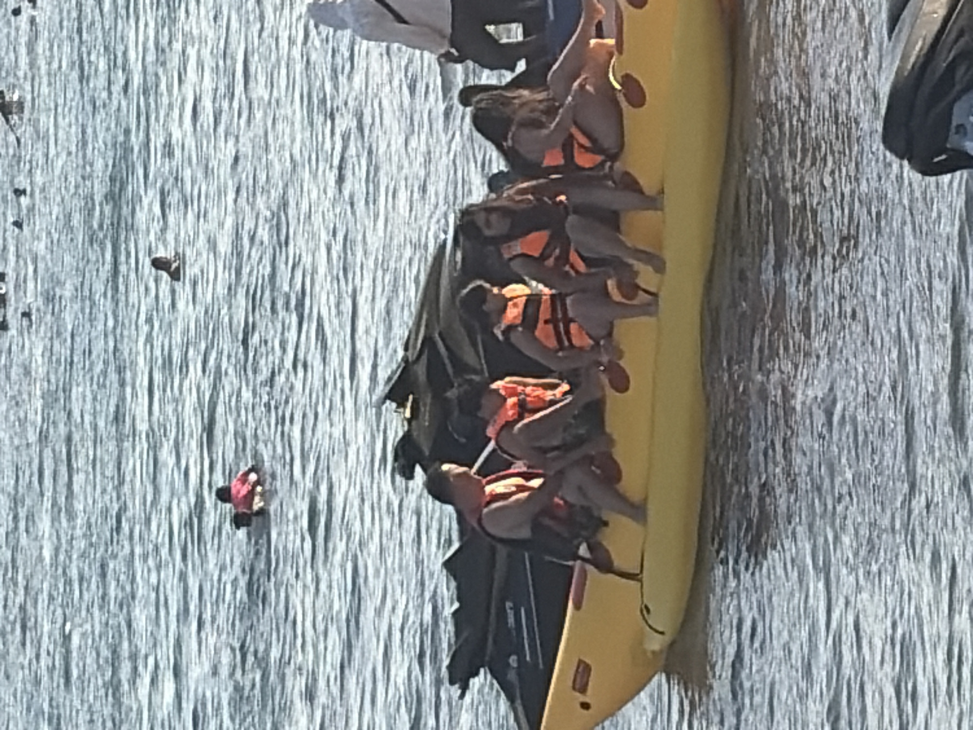 A group of people are on a yellow banana boat, some wearing orange life vests. The boat is in the water, with two jet skis behind it. In the background, other people are swimming in the water. The overall scene suggests a recreational activity in a body of water, likely a beach or resort area.
