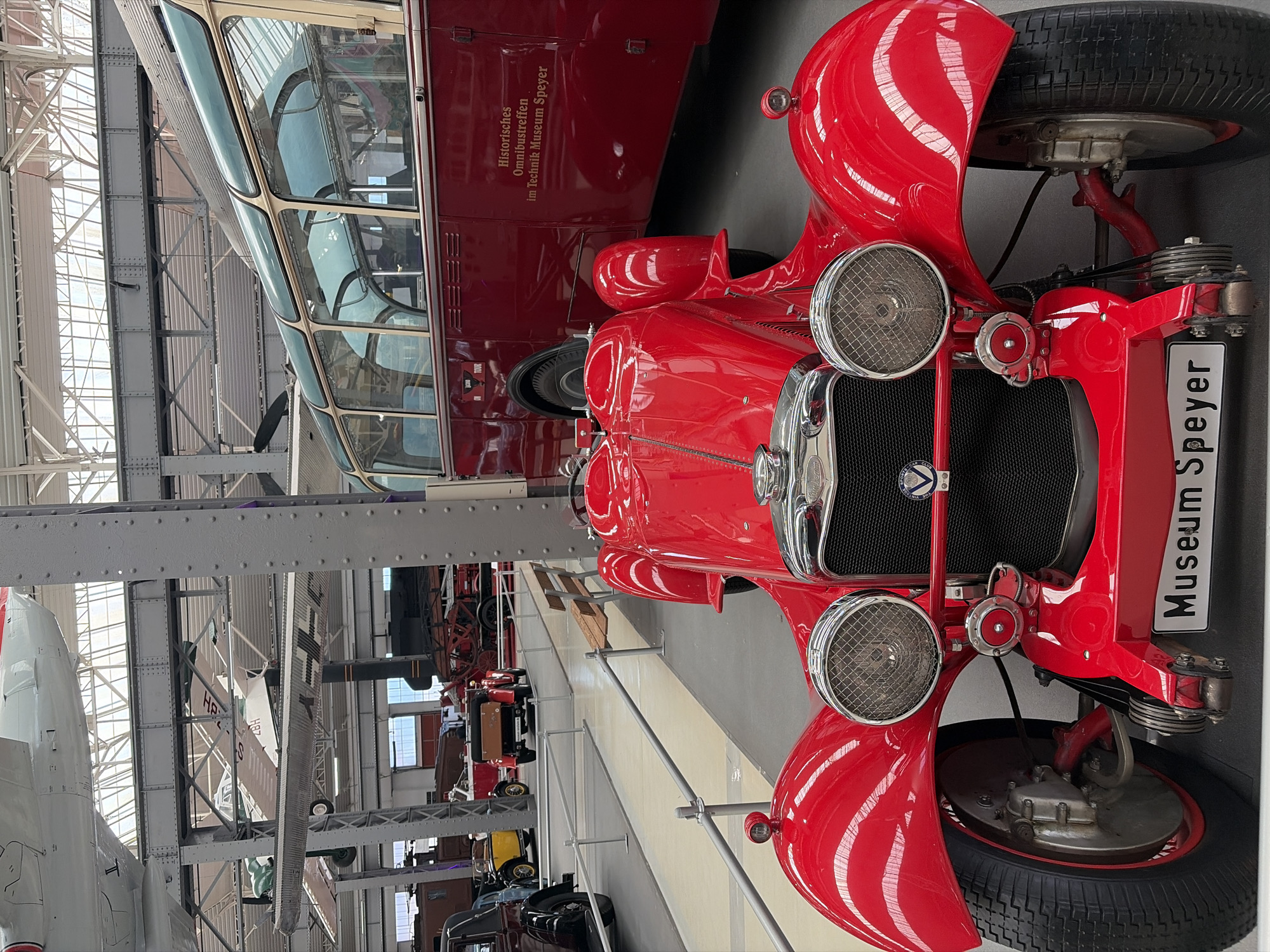 In a museum hall in Speyer, Germany, a bright red vintage car is displayed prominently. The car has a classic design with large, round headlights, a chrome grille, and exposed front wheels. A license plate on the front reads "Museum Speyer". Behind the car, a large, vintage red bus with a distinctive green-tinted roof is visible. The bus has text on its side that reads "Historisches Omnibustreffen im Technik Museum Speyer". The environment appears to be a spacious exhibition hall with exposed industrial ceilings and support beams, indicative of a museum setting. Several other exhibits, including what appear to be aircraft suspended from the ceiling and other vehicles in the background, can be seen. No people are visible in the frame.