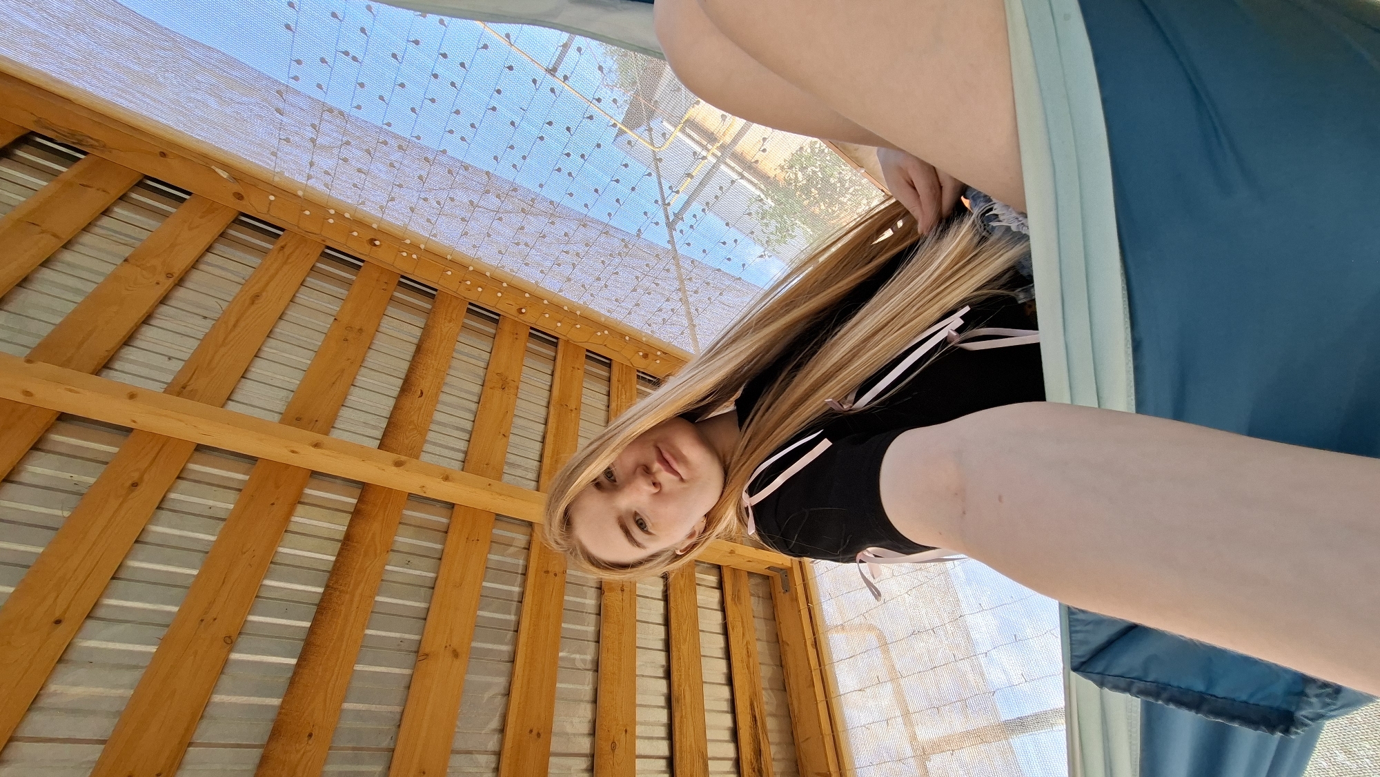 A low-angle shot depicts a young, fair-skinned woman with long blonde hair wearing a black top with pink accents. The setting appears to be a wooden gazebo or similar structure, as indicated by the wooden frame and roof above. The background reveals a partially obscured view of the sky through a patterned screen. The location is specified as Большой Букор, Russia.