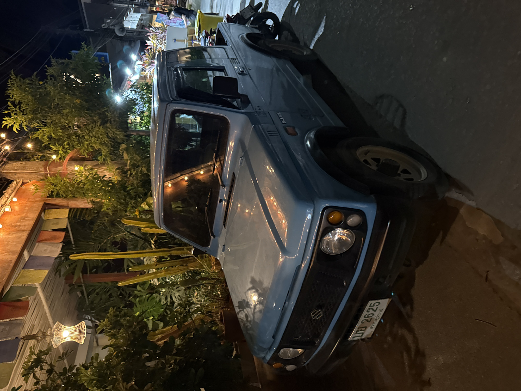 At night in the city of Baan Bo Phut, Thailand, a light blue Suzuki SJ410 utility vehicle is parked on a paved street. The vehicle has a hardtop cabin and an open bed in the rear. The license plate is visible and reads "UB 2625" followed by Thai script. The street is wet, indicating recent rain or cleaning.
In the background, there is a building with a covered porch decorated with prayer flags and string lights. Lush green plants, including cacti and large-leafed tropical foliage, are visible around the building, suggesting a garden setting. Power lines are strung across the dark night sky above. Further down the street, more buildings and lights can be seen, indicating a populated area. A person in dark clothing is standing near a yellow trash bin and a motorcycle parked behind the Suzuki. The lighting in the scene is a mix of artificial lights from the building, streetlights, and possibly interior vehicle lights, creating a somewhat dim and atmospheric environment.