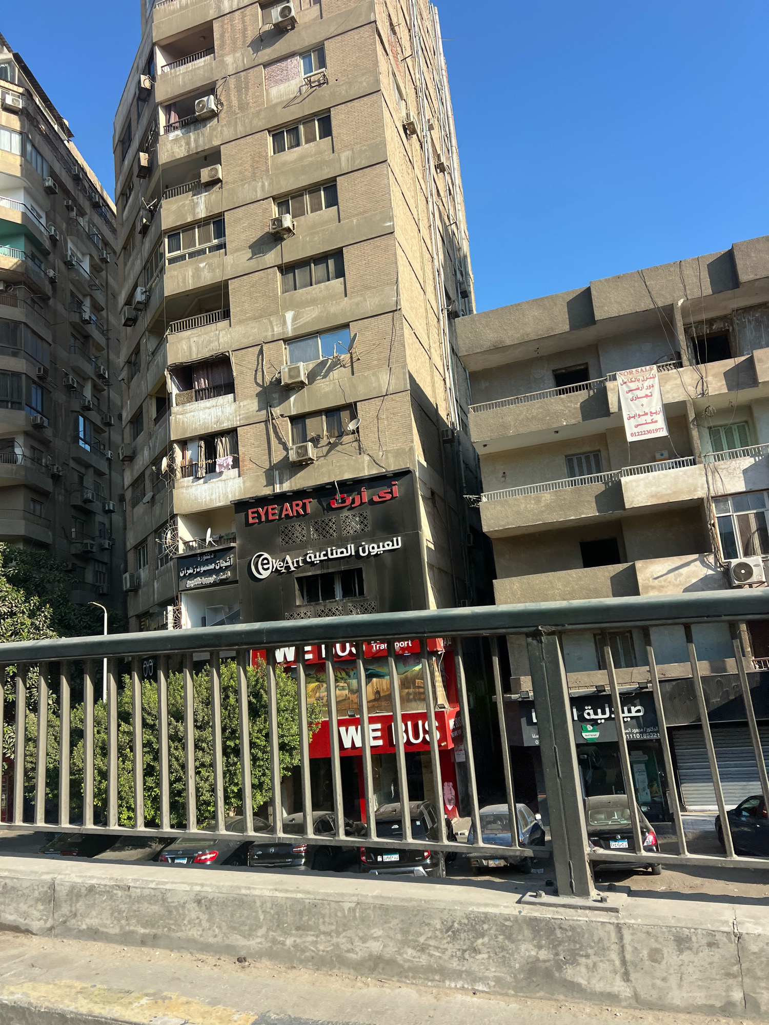 The image depicts a street scene in Giza, Egypt, focusing on a tall, weathered apartment building with numerous air conditioning units attached to its facade. The building is constructed from concrete and brick, showing signs of age and environmental wear. To the left, another similar building is partially visible, creating a dense urban environment.
At the base of the main building, several shops are present. One prominent sign reads "EYE ART" in English and Arabic. Another sign, also in Arabic, is for "العيون الصناعية EYE ART," which translates to "Artificial Eyes EYE ART." Below these, a red sign with white lettering says "WE BUS." Further to the right, another shop has Arabic lettering, and a "FOR SALE" sign in Arabic is affixed to an upper floor of a neighboring structure.
In front of the buildings, a metal fence runs horizontally across the foreground, partially obscuring the view of the ground level. Behind the fence, several parked cars are visible, including a dark-colored sedan with illuminated taillights and a lighter-colored vehicle. The sky above is clear and blue, indicating a sunny day. The overall impression is a bustling urban street with a mix of residential and commercial establishments.