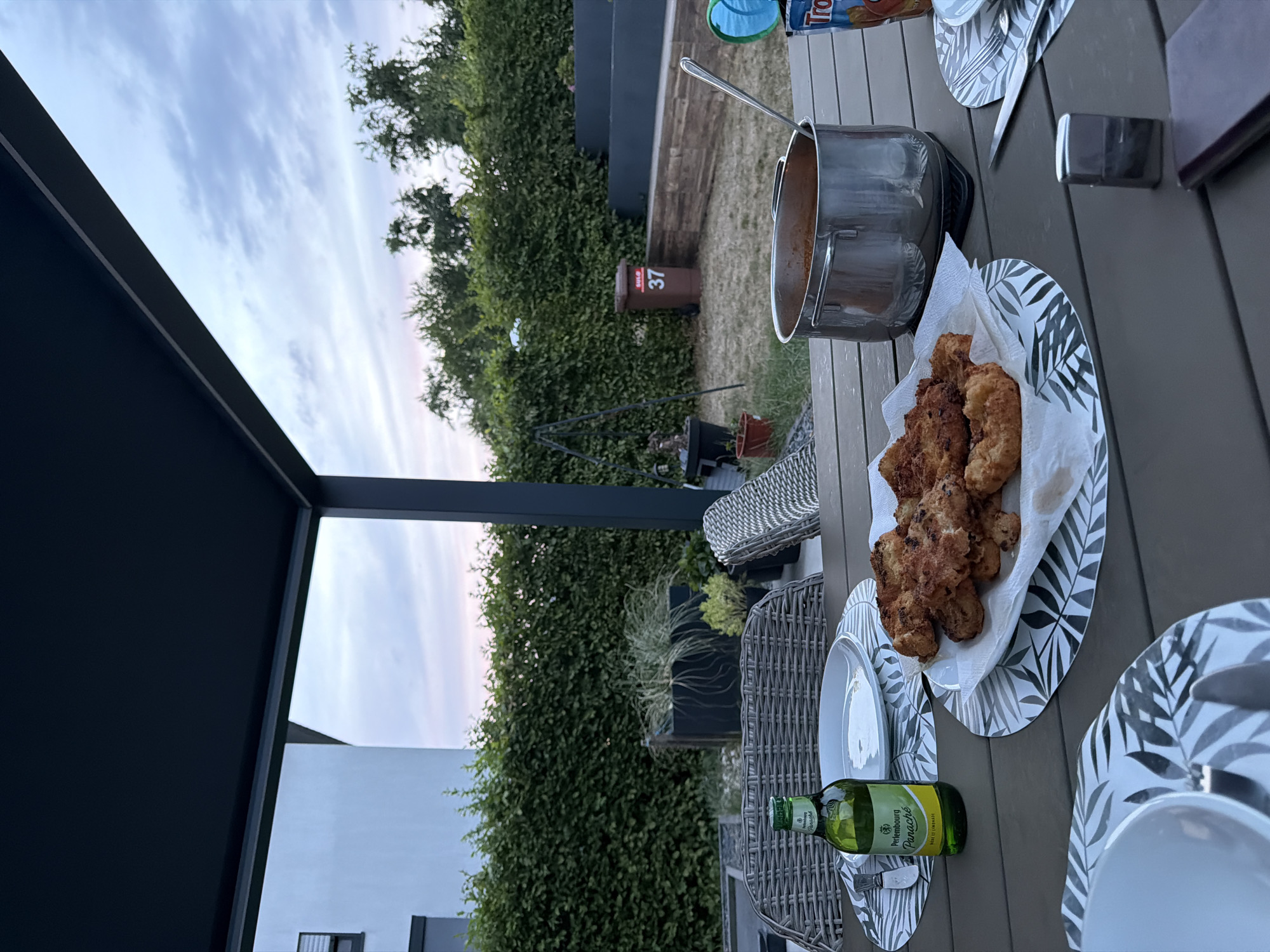 The image depicts an outdoor dining setting in Junglinster, Luxembourg, at dusk. A table is laden with food and drinks, including fried cutlets on a paper towel, a pot of sauce with a ladle, and a bottle of Perlenbourg Panaché. Plates and cutlery are also present. In the background, a neatly trimmed green hedge forms a backdrop, with trees beyond. A modern house with white walls and dark trim is visible to the left, and a brown trash bin with the number "37" is situated in the yard to the right. The sky shows hues of pink and blue, indicating the end of daylight. The scene appears to be a casual meal enjoyed on a patio or backyard.