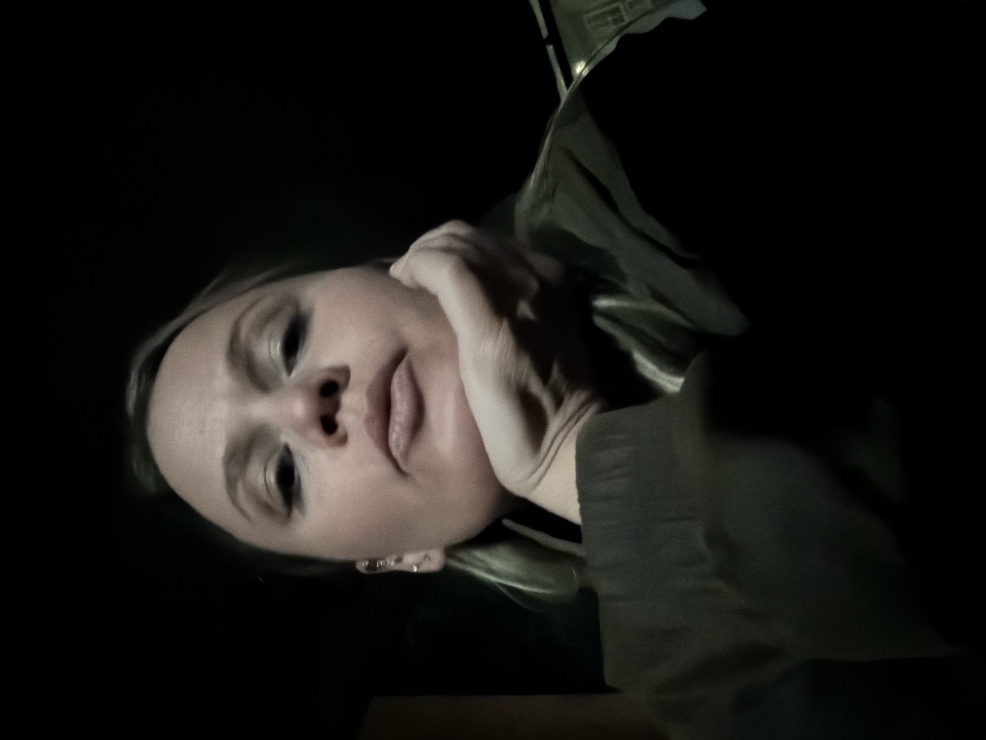 The image depicts an adult female in a dimly lit environment, purportedly in Dubnik, Russia. The subject, a fair-skinned individual with light-colored hair, occupies the majority of the frame, angled slightly from below, looking directly at the camera. Her right hand is observed with fingers curled, supporting her chin. She exhibits a neutral facial expression with slightly pursed lips. Multiple small metallic piercings are visible on her left ear. She wears a dark-colored garment, possibly a jacket or sweater. The background is almost entirely black, with a faint, indistinct structure or window outline barely perceptible in the lower right corner, suggesting artificial light or a distant reflective surface. The primary illumination appears to emanate from the front, highlighting the subject's face.