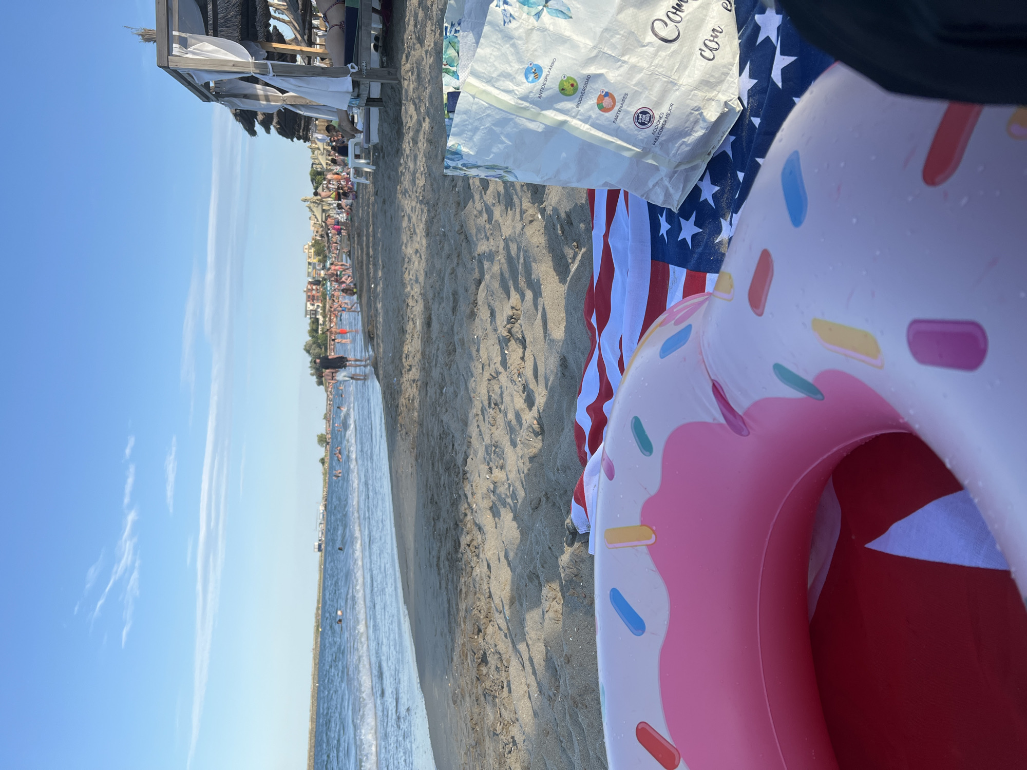 The image depicts a sunny day at a beach, likely in Constanța, Romania, given the context provided. In the foreground, a pink inflatable donut float with colorful sprinkles rests on a beach towel printed with the American flag. To the right, a white shopping bag with floral patterns and text in Spanish is placed on the towel. The sand is visible with some footprints. Beyond the immediate foreground, the sea stretches out, with gentle waves lapping the shore. Several people are visible in the water, swimming and enjoying the sea. Further inland, the beach is populated with numerous beachgoers relaxing on lounge chairs under thatched umbrellas. In the background, buildings with red roofs are visible along the coastline, characteristic of coastal towns. The sky is clear and blue, with a few wispy clouds, indicating good weather. The overall scene conveys a relaxed and vibrant atmosphere of a busy beach resort.