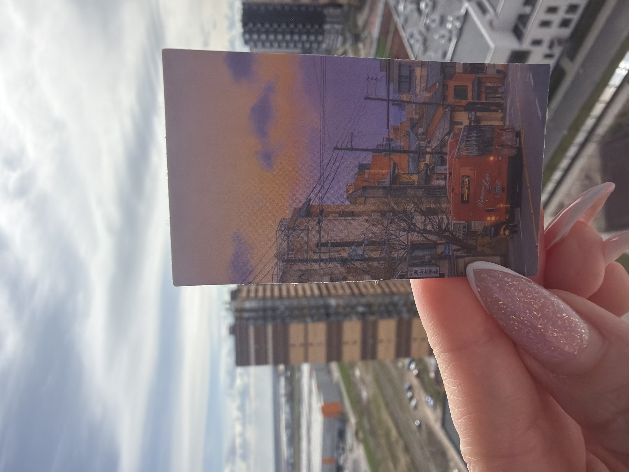 The image displays a hand holding a small, rectangular print or postcard against a backdrop of an urban landscape, captured from a high vantage point in Saint Petersburg, Russia. The entire photo is rotated 90 degrees clockwise, presenting the real-world horizon on the right side.
A human hand, located in the lower right portion of the frame, holds the postcard horizontally. The person's nails are long, pointed, and feature a glittery pink polish with distinct white French tips.
The postcard itself depicts an atmospheric street scene, likely at sunset or twilight, with a sky transitioning from warm orange to soft purple hues. The illustration features dense utility poles and an intricate network of power lines, characteristic of some East Asian cityscapes. Numerous buildings line the street, and a prominent red bus with visible headlights and a white roof is positioned on the right side of the postcard's scene. Faint English text is discernible on the side of the bus, possibly reading "Yokohama" and "Bus." Below the bus, on a small sign, Japanese characters are visible, faintly spelling "ヨコハ" (YOKOHA). A tree with bare branches stands in the foreground of the postcard's illustration.
The background of the overall photo reveals a sprawling urban environment under an overcast sky. Tall, multi-story buildings with repetitive window designs extend upwards from below, indicative of a densely built-up city. Far below, a wide street or open area with parked vehicles can be seen. The real-world sky is bright and filled with soft, diffused clouds.
