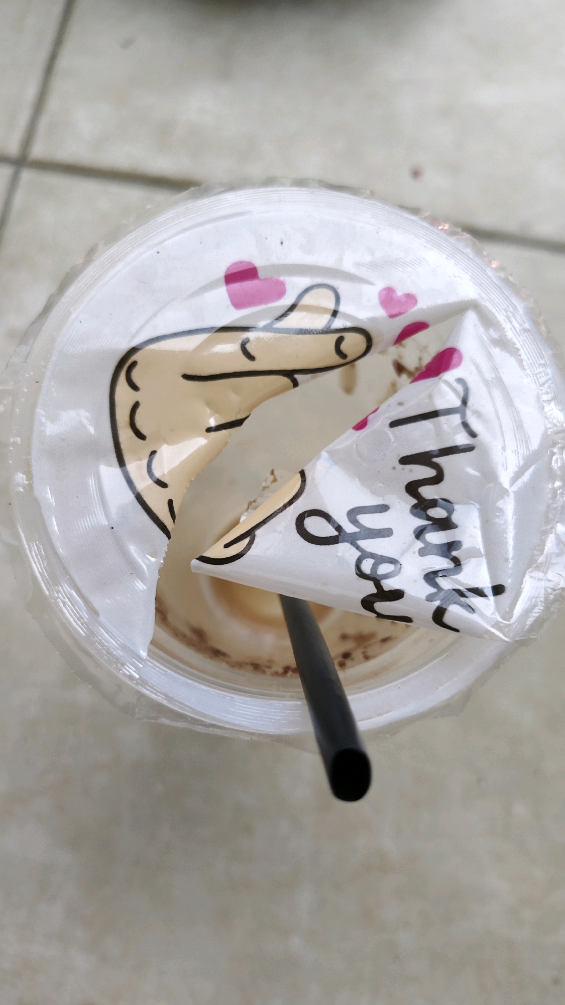 A top-down, close-up shot captures a clear plastic cup filled with a light brown beverage, likely coffee or a similar drink, with a black straw protruding from the center. The cup's lid is partially peeled back, revealing a printed design of a cartoon hand giving a thumbs up and the words "Thank you" written in cursive. Pink hearts are also visible on the lid. The cup sits on a light-colored tiled surface, possibly a floor or table, in what appears to be an outdoor or semi-outdoor setting. There is no visible indication of the specific location within Pangpajung, Indonesia, other than the tiled flooring which is common in many establishments.