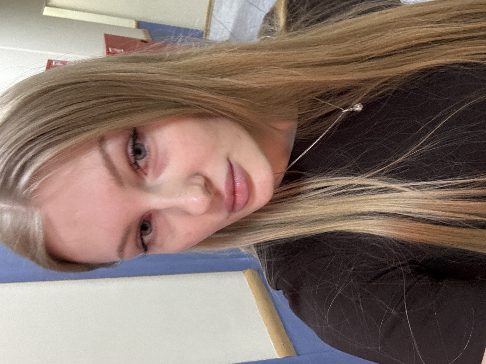A close-up portrait shows a young woman with light blonde hair, wearing a dark-colored top and a delicate silver necklace. The background reveals glimpses of an interior with blue and white surfaces. The image does not provide sufficient detail to identify a specific location in Perm, Russia, or any explicit actions or interactions.