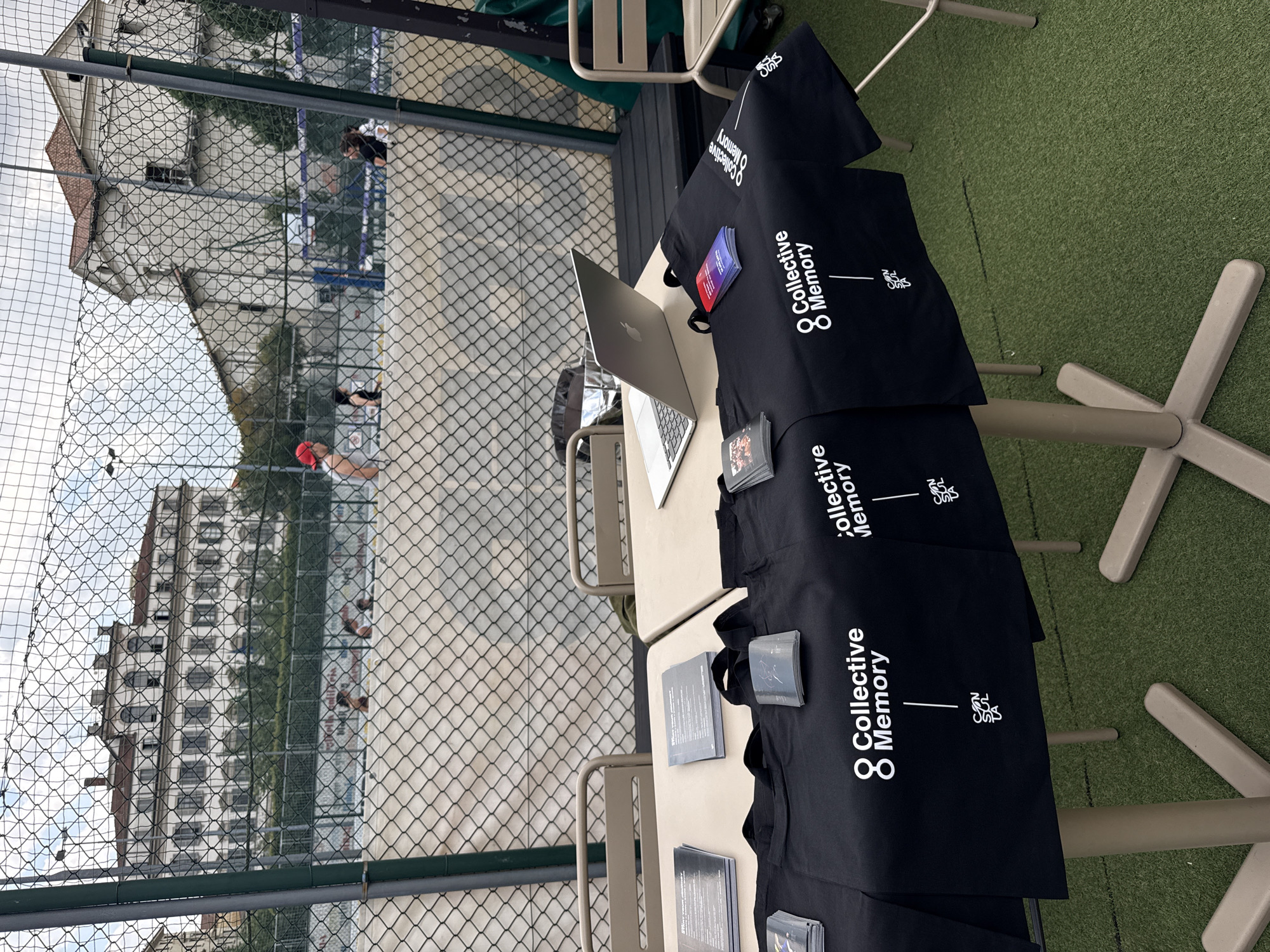 The image captures an outdoor scene at a sports facility in Milan, Italy. In the foreground, two light-colored rectangular tables are set up on artificial green turf. The tables are draped with black fabric covers displaying white text logos: "Collective Memory" (with an '8' symbol) and "CEN SKOL." On the left table, multiple stacks of dark-colored brochures or booklets, some titled "START YOUR JOURNEY," are arranged, alongside smaller stacks of square-shaped items. On the right table, an open silver-colored laptop is present, flanked by additional stacks of brochures, some featuring colorful gradient covers. Several black tote bags, branded with the same logos, are displayed across both tables. Two light-colored metal chairs are visible behind the tables.
Separating the foreground from the midground is a chain-link fence with dark green vertical supports. Beyond this fence, outdoor sports courts, consistent with padel or tennis courts, are visible. Four individuals are present on the courts: two on the left court and two on the right court. One individual on the right court is wearing a red cap. Additional chain-link fencing and court walls separate the individual playing areas.
In the background, behind the courts, stand several large, light-colored buildings of classical European architecture. The most prominent building on the left is a multi-story structure with a tiled roof, numerous windows (some arched), and decorative architectural details. Green foliage is visible between the courts and the buildings. The sky above is partially cloudy.