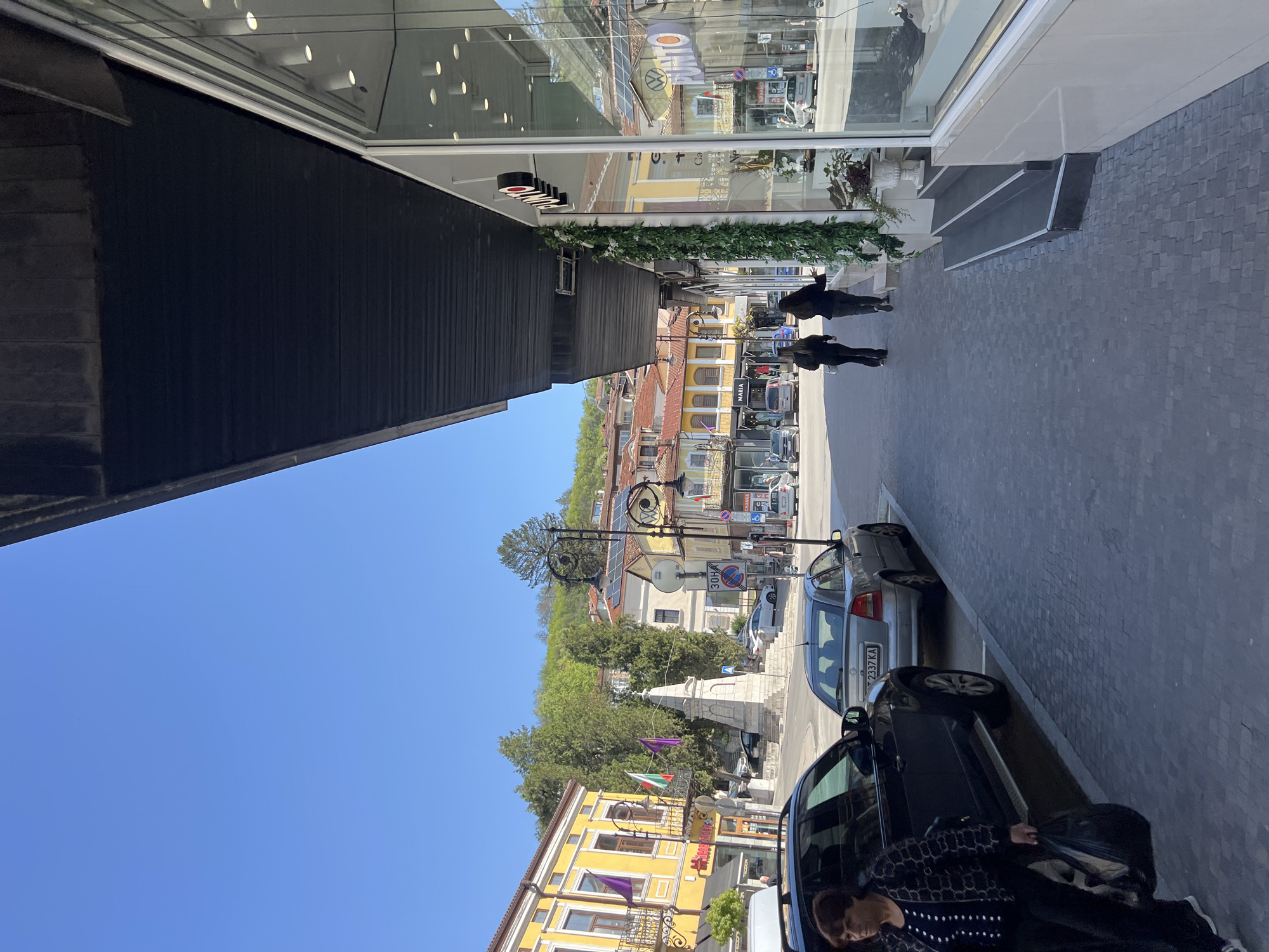 The image presents a vibrant street scene in Veliko Tarnovo, Bulgaria, captured on a clear and sunny day with a bright blue sky. The perspective looks down a street that blends modern and traditional architecture.
On the left, a contemporary building features a large, reflective glass facade, mirroring elements from across the street such as older buildings, parked cars, and the clear sky. Below the windows, a dark, ribbed paneling covers the lower part of the building, adorned with a strip of artificial greenery. Reflections in the windows display visible text like "SUSHI" (reversed) and a "VOLKSWAGEN" logo, along with interior ceiling lights.
The right side of the street is lined with colorful, multi-story buildings, many featuring traditional yellow plaster and red-tiled roofs, indicative of Bulgarian architecture. The street itself is paved with dark, patterned cobblestones or pavers. Several cars are parked along the curb, including a silver sedan with a Bulgarian license plate "2337 KA" and a dark car in the foreground. A partially visible person is seen near the dark car, while two other individuals walk further down the street.
Further into the scene, a decorative black street lamp post and a stone monument or fountain are visible. Flags, including one with the white, green, and red stripes of the Bulgarian flag, hang near the monument. Traffic signs are present, including one indicating a "30KM" speed limit and another with Cyrillic text, possibly "МАРИЯ" (Maria). The background features lush green hills, completing the urban landscape. The overall scene suggests a pleasant day for pedestrians and everyday city life.