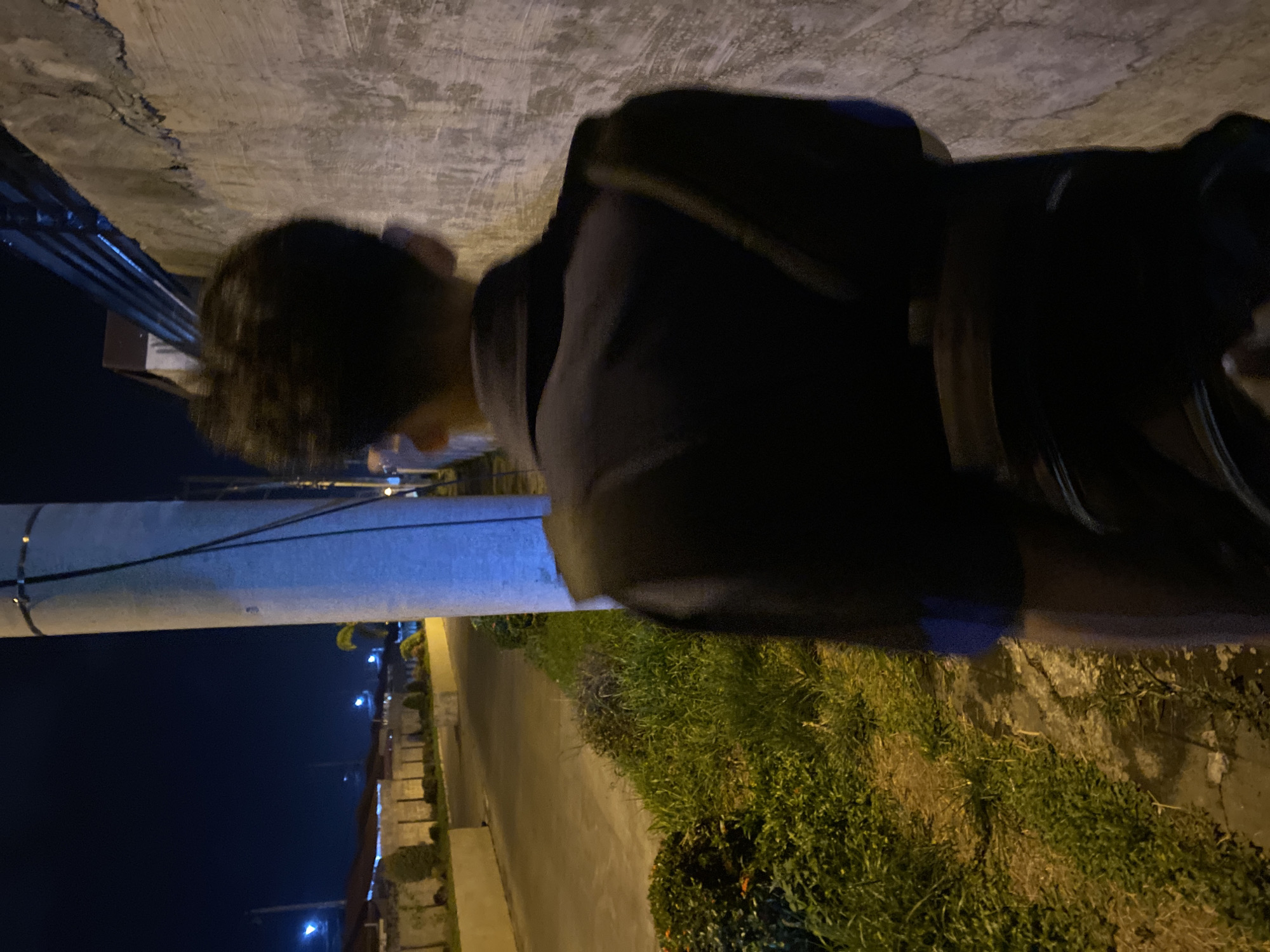 A nighttime image captures the rear perspective of an individual walking along a paved path in Davao City, Philippines. The subject, a person with dark, short hair, is viewed from behind and slightly to the left, wearing a dark long-sleeved garment with a blue accent on the right arm. A dark strap from a bag is visible over the right shoulder, and another dark strap encircles the lower back.
On the left, a narrow paved road or path is bordered by grass and low bushes. In the distant background, streetlights emit blue-white light, illuminating residential buildings with sloped roofs and concrete perimeter walls. Concrete planters are visible along the road's edge. To the right, a textured, light-grey concrete wall shows cracks and dark streaks. Above the wall, a metal fence is discernible. Centrally positioned, a large, light-colored utility pole features dark cables running vertically. Artificial light sources illuminate the scene, highlighting the pole and wall, and casting shadows across the foreground vegetation. The overall environment is consistent with a quiet urban or suburban street at night.
