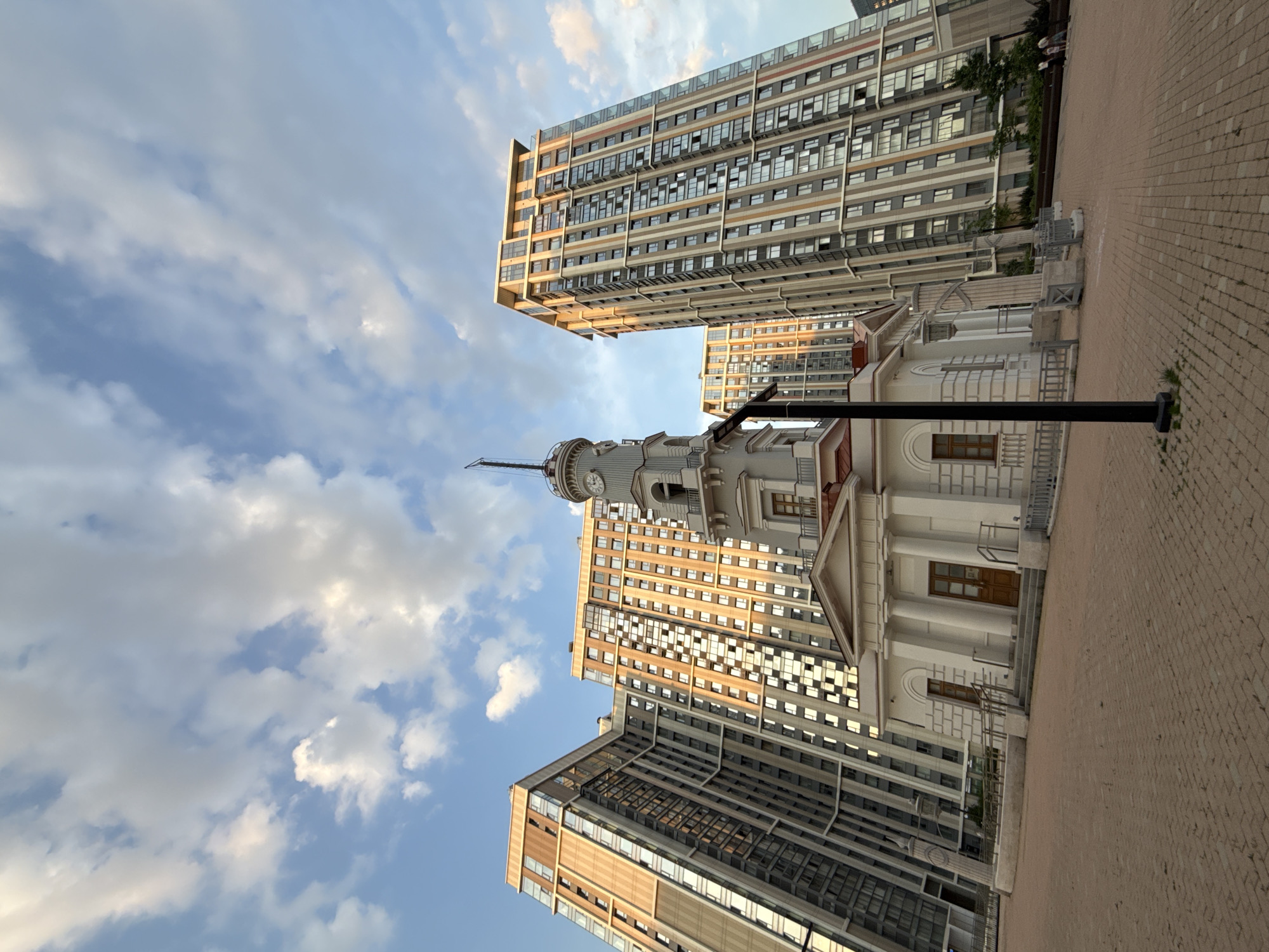 The image shows a street scene in Moscow, Russia. A white, classical-style building with a clock tower stands in the foreground, positioned between two modern high-rise buildings. The sky is visible with white clouds and patches of blue. A black lamppost is present near the white building. The ground is paved with bricks. The perspective is angled upwards, emphasizing the height of the buildings and the sky. No people are clearly visible.