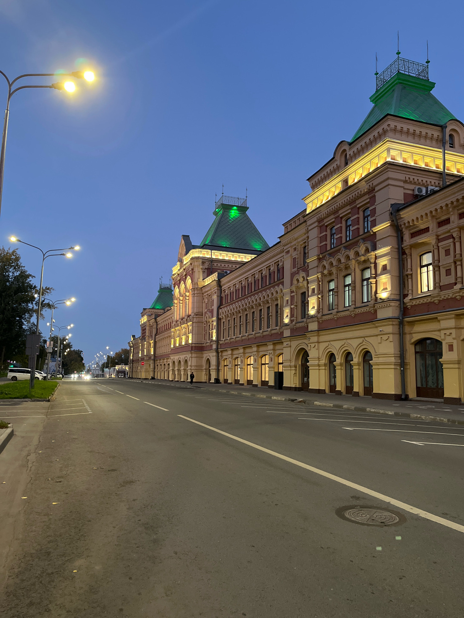 This is a medium-angle, eye-level, outdoor, full-shot photograph of a street in Nizhny Novgorod, Russia, taken at dusk. On the right is a large, ornate building with a red and cream facade, arched windows, and green roofs, illuminated by exterior lights. The building appears to be a historical architectural landmark. Across the street on the left, several cars are parked along the sidewalk, and a few more vehicles are driving on the road. Streetlights with multiple lamps are visible, casting a yellow glow. The sky is a clear, deep blue, transitioning into twilight. The street is paved and lined with white markings, and there is a manhole cover visible in the foreground. Trees are present on the far left side of the image. The scene depicts a quiet urban environment as night begins to fall.