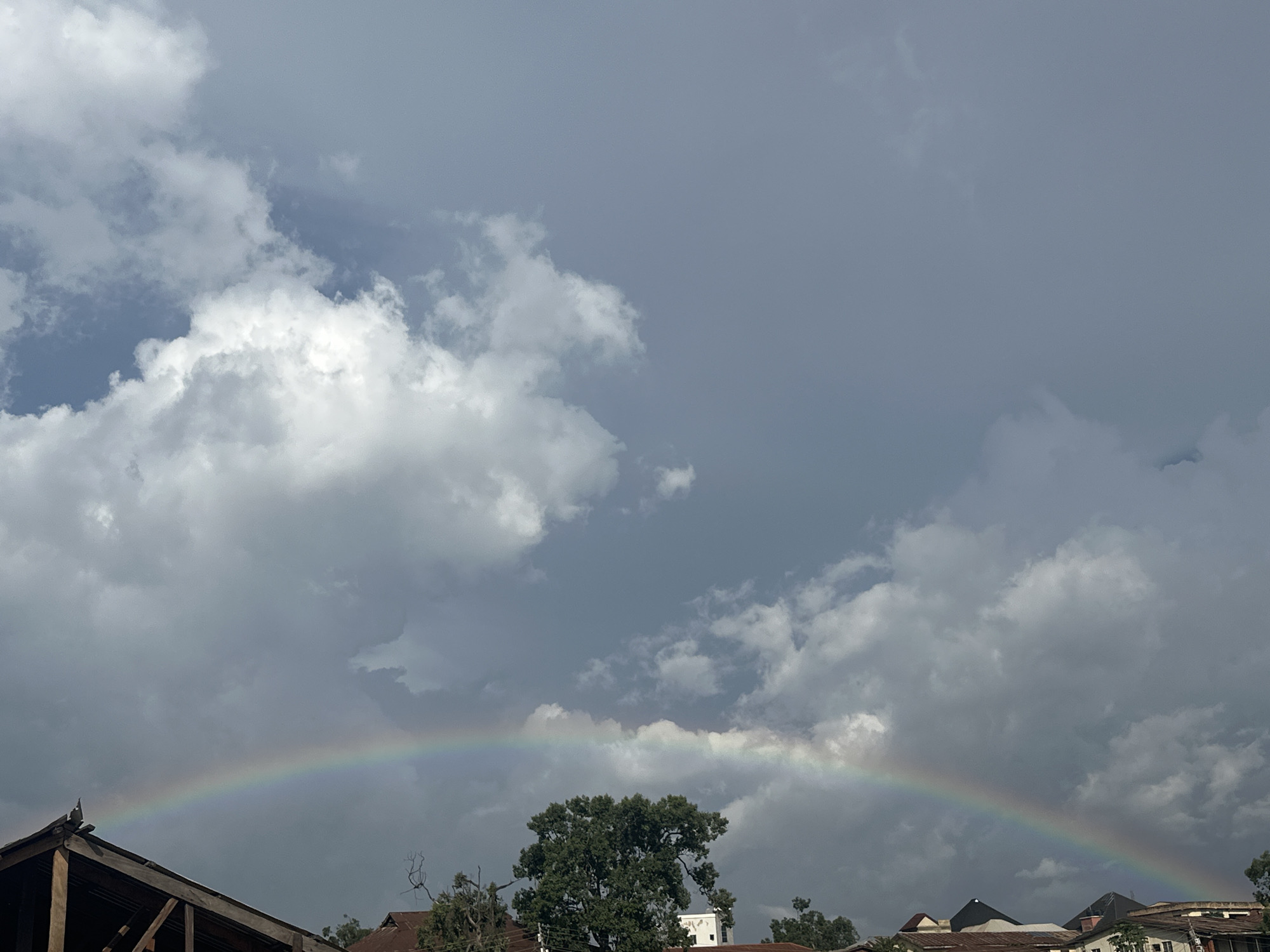 The image depicts a view of the sky above Jos, Nigeria, featuring a rainbow arching across the frame. The sky is predominantly filled with dramatic, gray clouds, interspersed with patches of brighter, white clouds. The lower portion of the image shows rooftops of buildings, some with corrugated iron roofs, and the upper branches of trees. A large, leafy tree stands prominently in the mid-ground. The rainbow is visible in the lower half of the sky, its colors muted by the overcast conditions. The overall scene suggests a post-rain or intermittent rain scenario in an urban environment.
