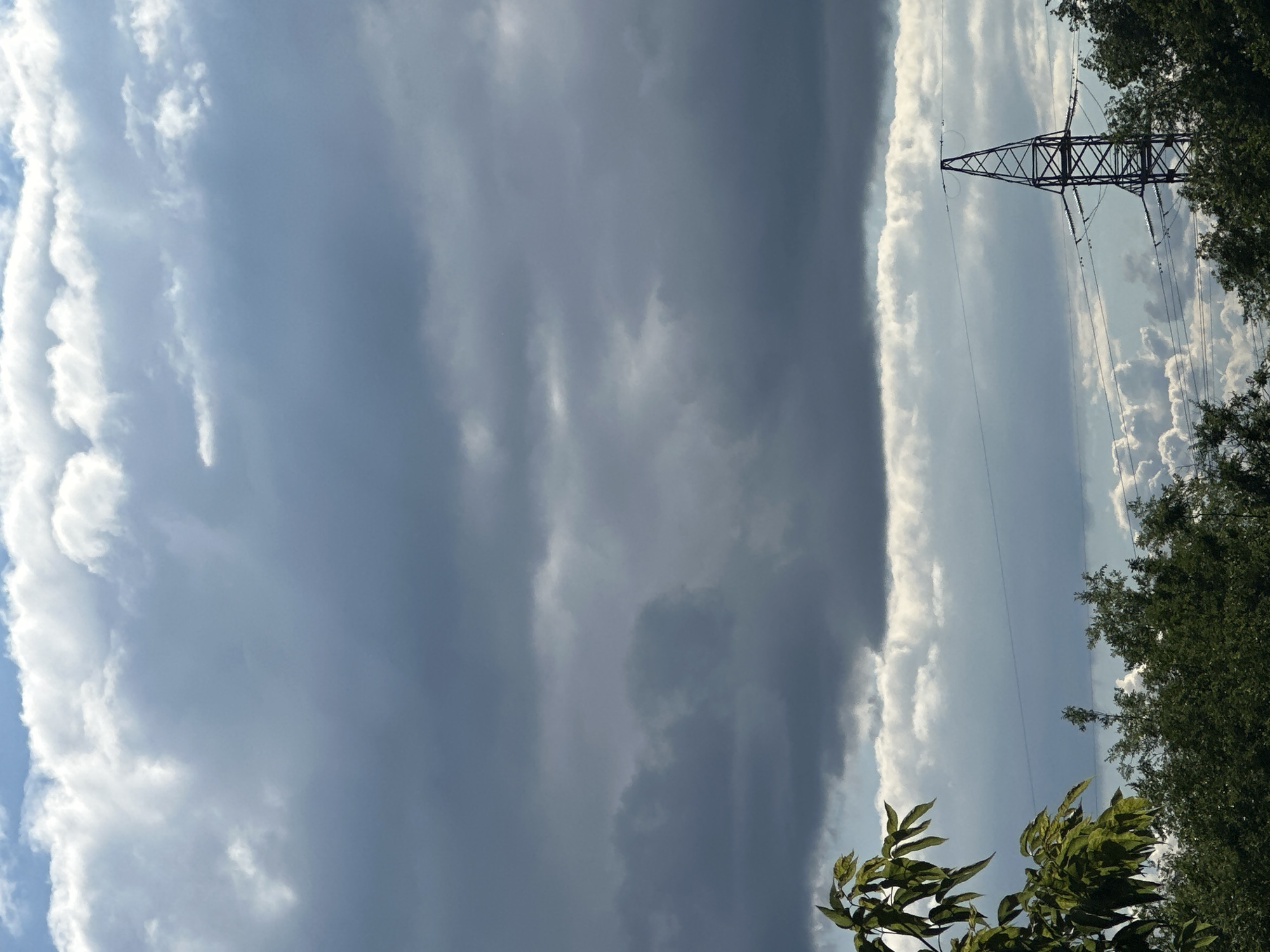 The image shows a wide shot of a cloudy sky, with a power line tower in the background and trees on the right. The clouds are varied, with some appearing dark and heavy, possibly indicating an approaching storm, while others are bright and white, suggesting sunshine breaking through. A tall, lattice-style power line tower stands prominently in the middle ground, with power lines stretching across the sky. Lush green trees frame the right side of the image, their leaves casting shadows. The scene appears to be outdoors, likely during the daytime, given the lighting. The overall impression is one of a dynamic sky with a mix of light and dark clouds, set against a natural landscape with man-made infrastructure.