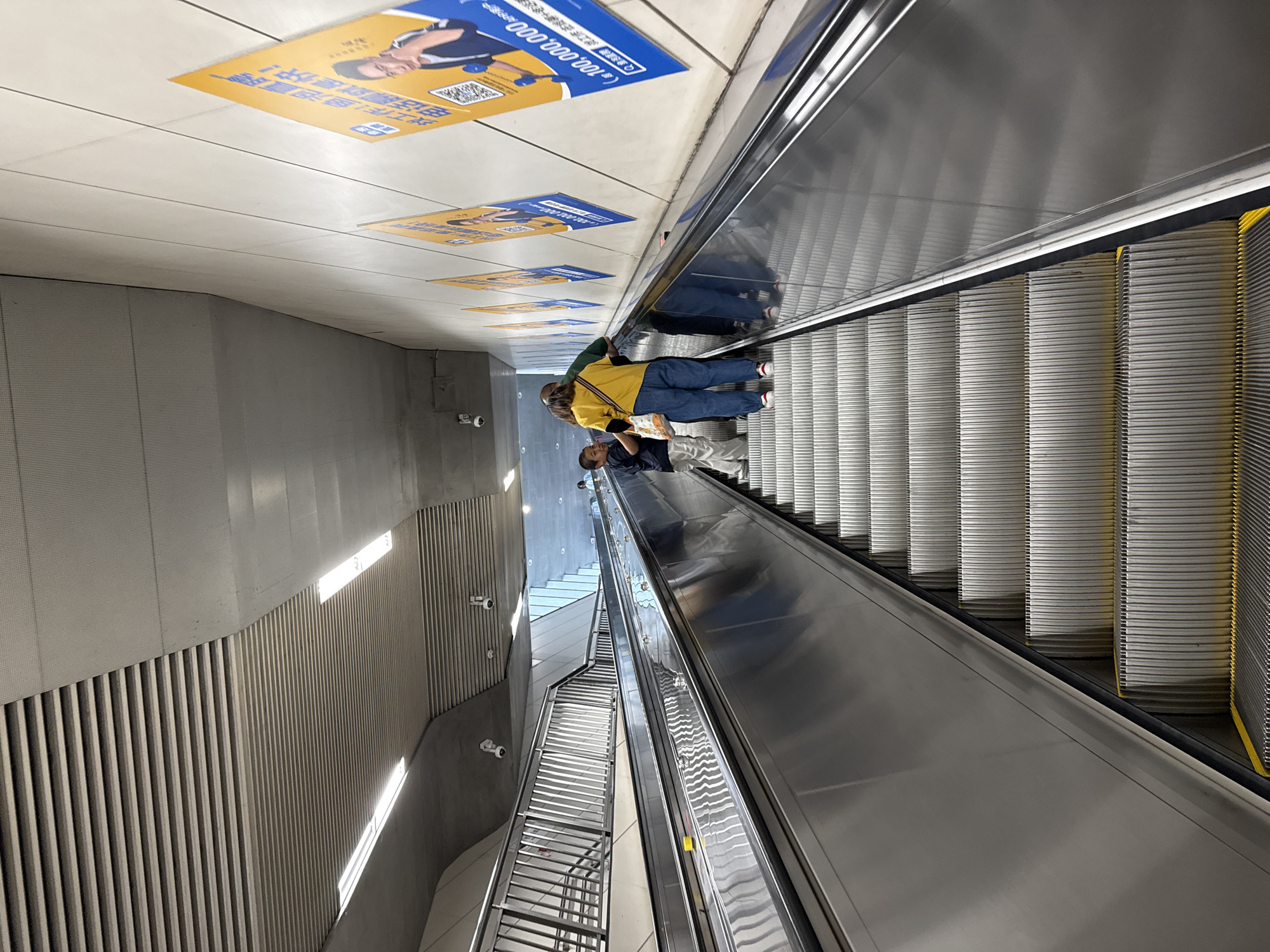 This image captures an upward-moving escalator system within a public transit area, likely a subway station, located in Haidian District, China.
Two parallel escalators are featured, with the one on the right occupied by three individuals ascending. The lead person, an adult, wears a yellow short-sleeved top, blue jeans, and red-soled shoes, carrying a light-colored shoulder bag. Immediately behind this individual is another adult wearing a dark green short-sleeved top. To the left of the yellow-shirted adult and slightly in front, a child dressed in a dark t-shirt and light-colored pants stands, appearing to look towards the yellow-shirted adult. Reflections of these three individuals are visible on the polished metal side panel of the right escalator.
The escalators are constructed with silver-colored ribbed steps, clearly delineated by yellow lines at the front edge of each step, and feature black handrails. A small, yellow, rectangular object rests on the central divider between the two escalators.
The right wall is a light gray or white color, paneled with subtle horizontal lines. Multiple yellow and blue rectangular posters are affixed to this wall, each featuring a male figure, Chinese text, and a QR code; one prominent poster displays "Q-100,000,000". The ceiling is multi-tiered, incorporating both slatted gray panels and smooth light gray sections. Several recessed linear light fixtures emit bright white illumination. Multiple white dome-shaped security cameras are mounted on the ceiling and upper wall sections. On the left side of the scene, a multi-bar stainless steel railing system runs parallel to the escalators, separating them from an adjacent area which also appears to contain another set of stairs further back.