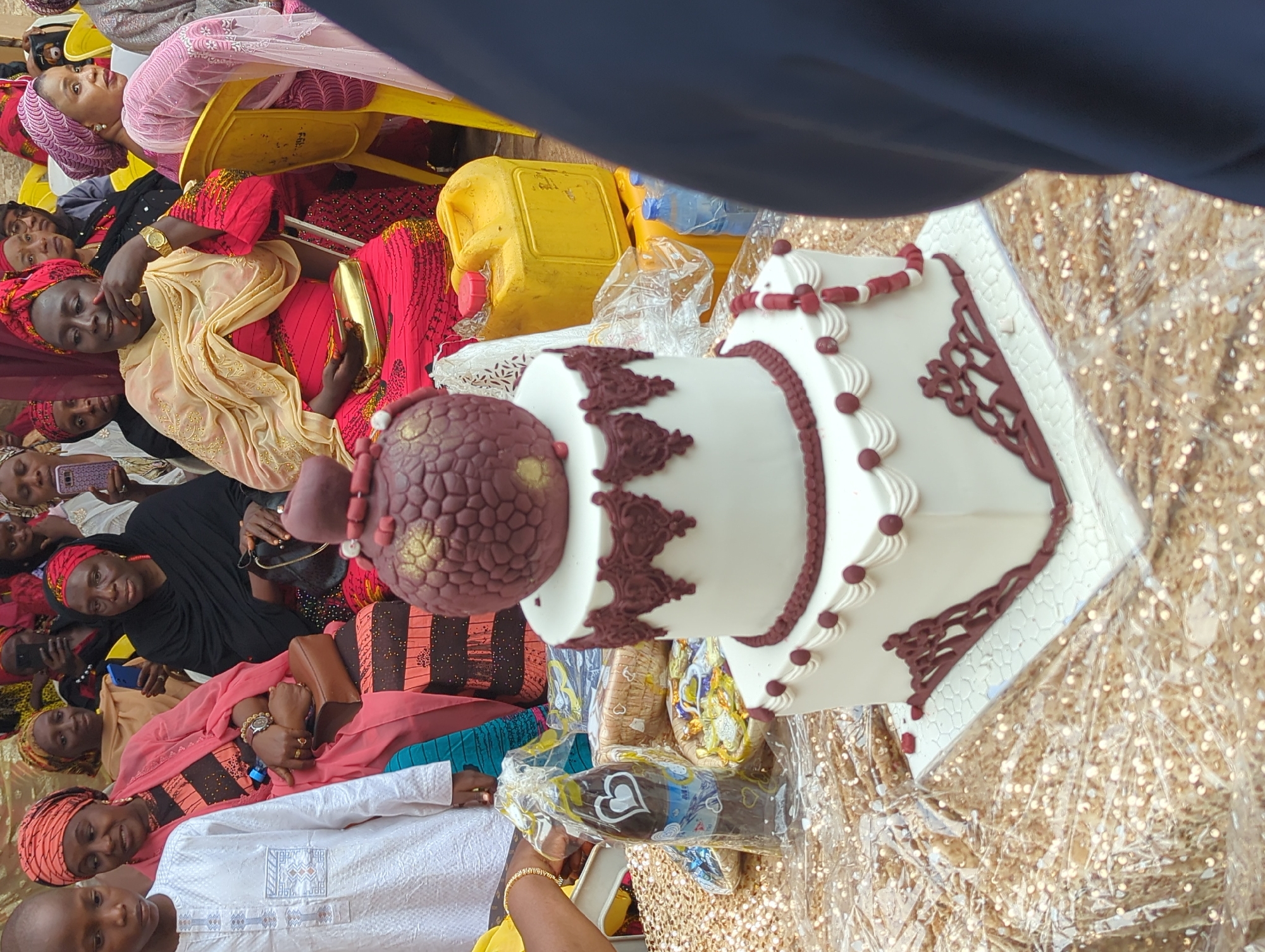 This image, captured in Unguwar Rimi, Nigeria, displays an outdoor social gathering with a prominent two-tiered cake as the central subject. The cake is positioned on a table covered with gold sequined fabric and a clear plastic sheet. It features white frosting with maroon-brown decorative details; the upper tier has a scalloped border, while the lower tier exhibits a beaded string and intricate scrollwork at its base. A dark reddish-brown ovoid cake topper with gold accents and maroon beads rests on the top tier.
To the left of the cake, a plastic-wrapped bottle with a heart emblem and partially visible "Mr." label is present, along with a woven basket containing small, individually wrapped items. In the background, behind the cake and to its right, two yellow plastic jerrycans are stacked, with several blue plastic water bottles visible below.
A group of individuals is seated in the background. From left to right: a young boy in a white traditional garment with blue embroidery looks slightly right; a woman wearing a salmon-colored headwrap and a striped dress holds a brown purse and displays a gold watch on her left wrist; another woman with a yellow headwrap is partially visible; a woman in a red headwrap and dark clothing holds a dark purse; a woman in a red dress with a cream head covering, also wearing a gold watch and holding a gold clutch, rests her chin on her left hand; and a woman in a pink headwrap and pink garment is seated in a yellow plastic chair. All individuals are dressed in traditional West African attire.