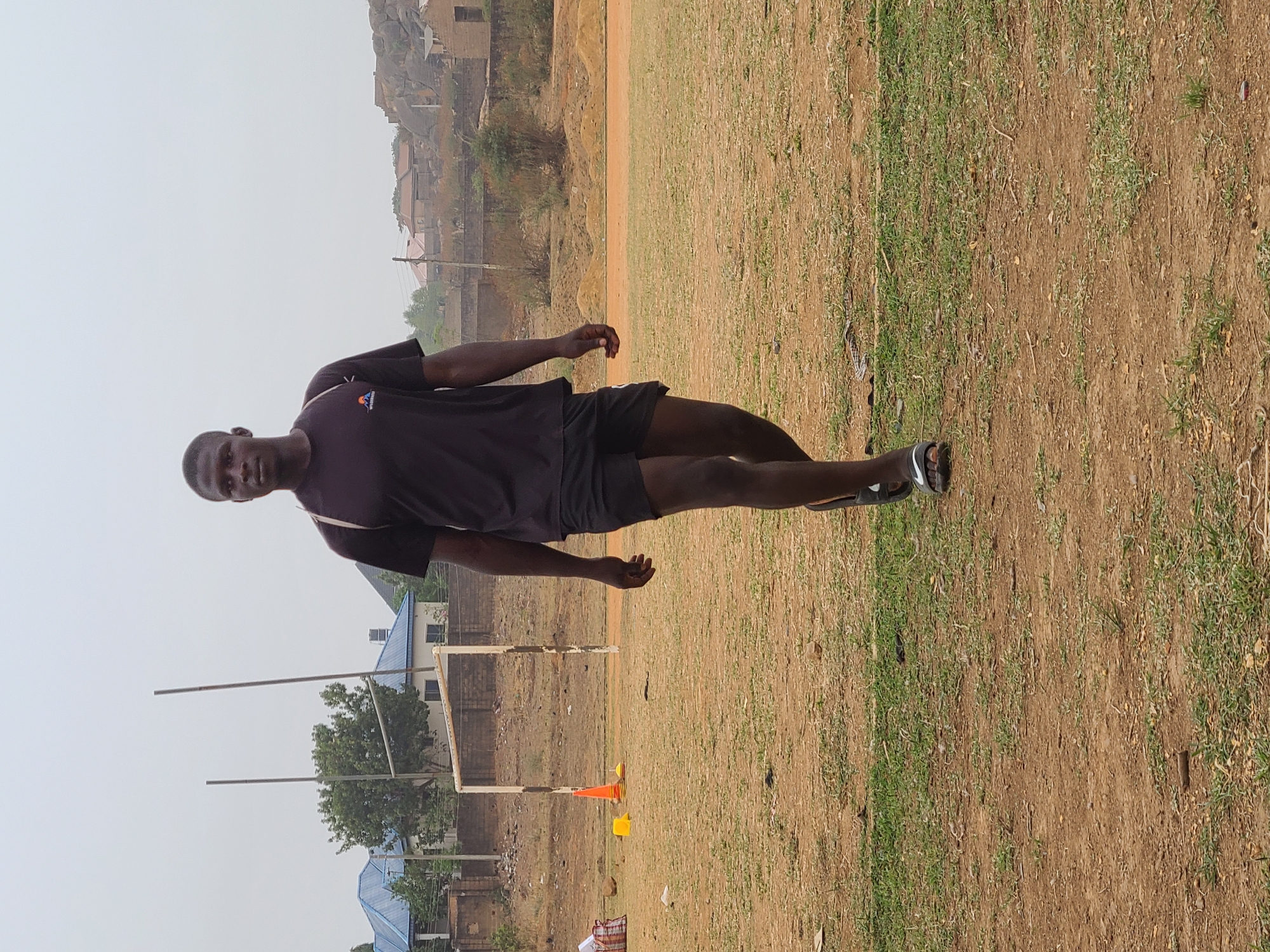 The image shows a man standing on a grassy field in Barikin Jirga Summa, Nigeria. He is wearing a dark-colored t-shirt, black shorts, and sandals. In the background, there is a dirt track, houses, a soccer goal with no net, and some training cones. The sky is overcast.