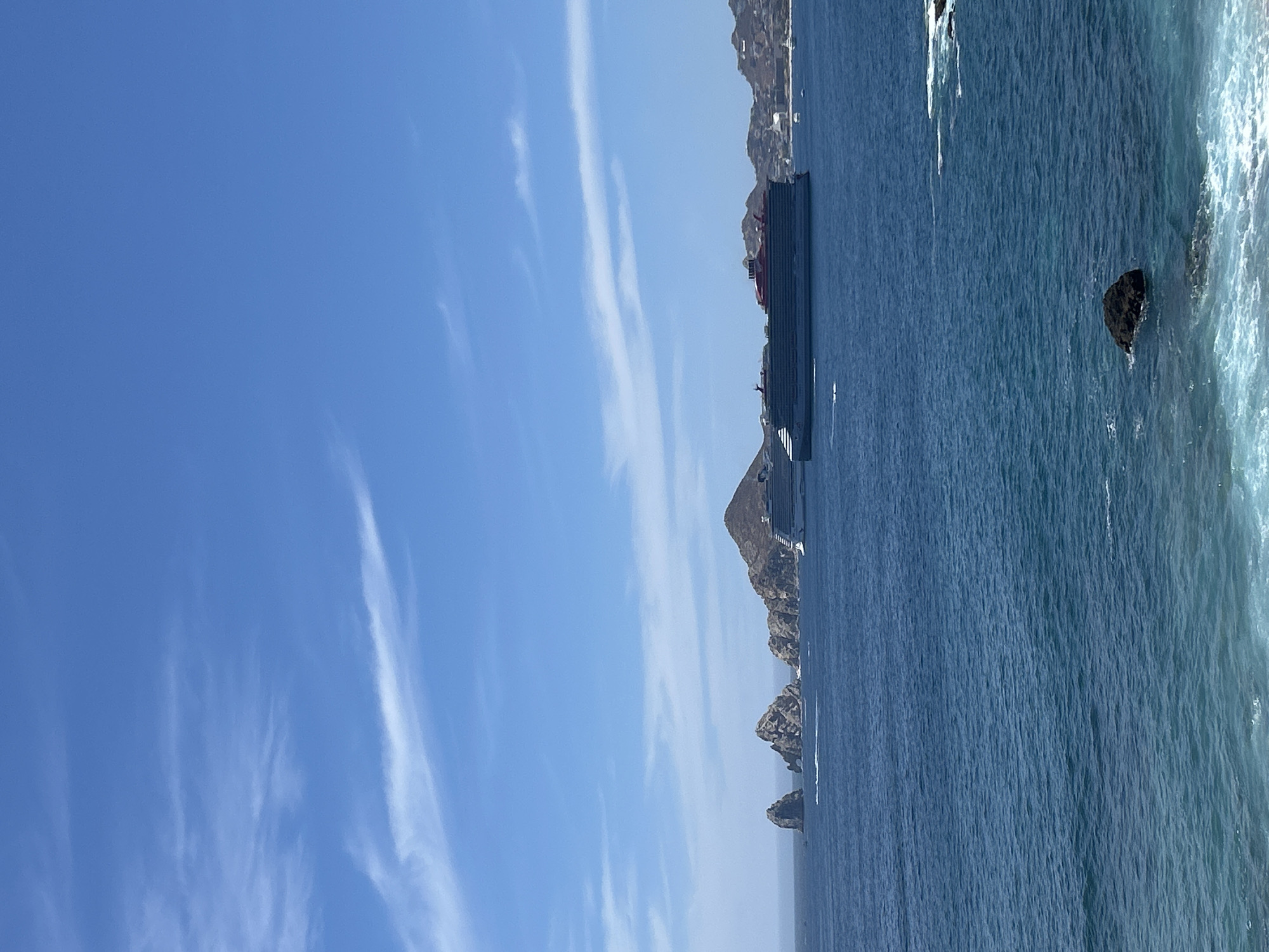 Captured from a vertical perspective, the image features a seascape likely located in Cabo San Lucas, Mexico. The scene is divided between a clear blue sky with high, wispy clouds and the deep blue expanse of the ocean. On the right side of the image, rocky cliffs and formations jut from the sea, defining the coastline. A large cruise ship is anchored off the coast, appearing between the cliffs and the open ocean. The water shows some signs of waves crashing against the shore, creating a foamy texture. The overall atmosphere is bright and sunny, typical of a coastal region.