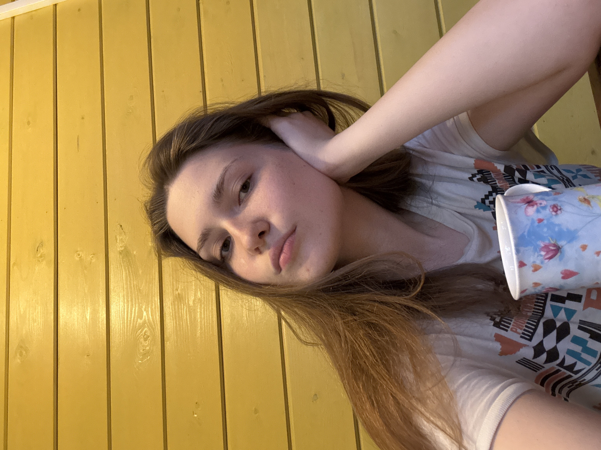 A young woman with long brown hair and fair skin is pictured in a medium close-up shot. She is facing the camera at an angle, with her left hand behind her head and her right arm resting across her body. She is wearing a white t-shirt with a colorful geometric pattern. In her right hand, she holds a mug with a light blue background and floral designs.
The background is a yellow wooden wall with vertical planks. The scene appears to be indoors. The location is not clearly discernible from the image and cannot be determined as Ленинградская, Russia.