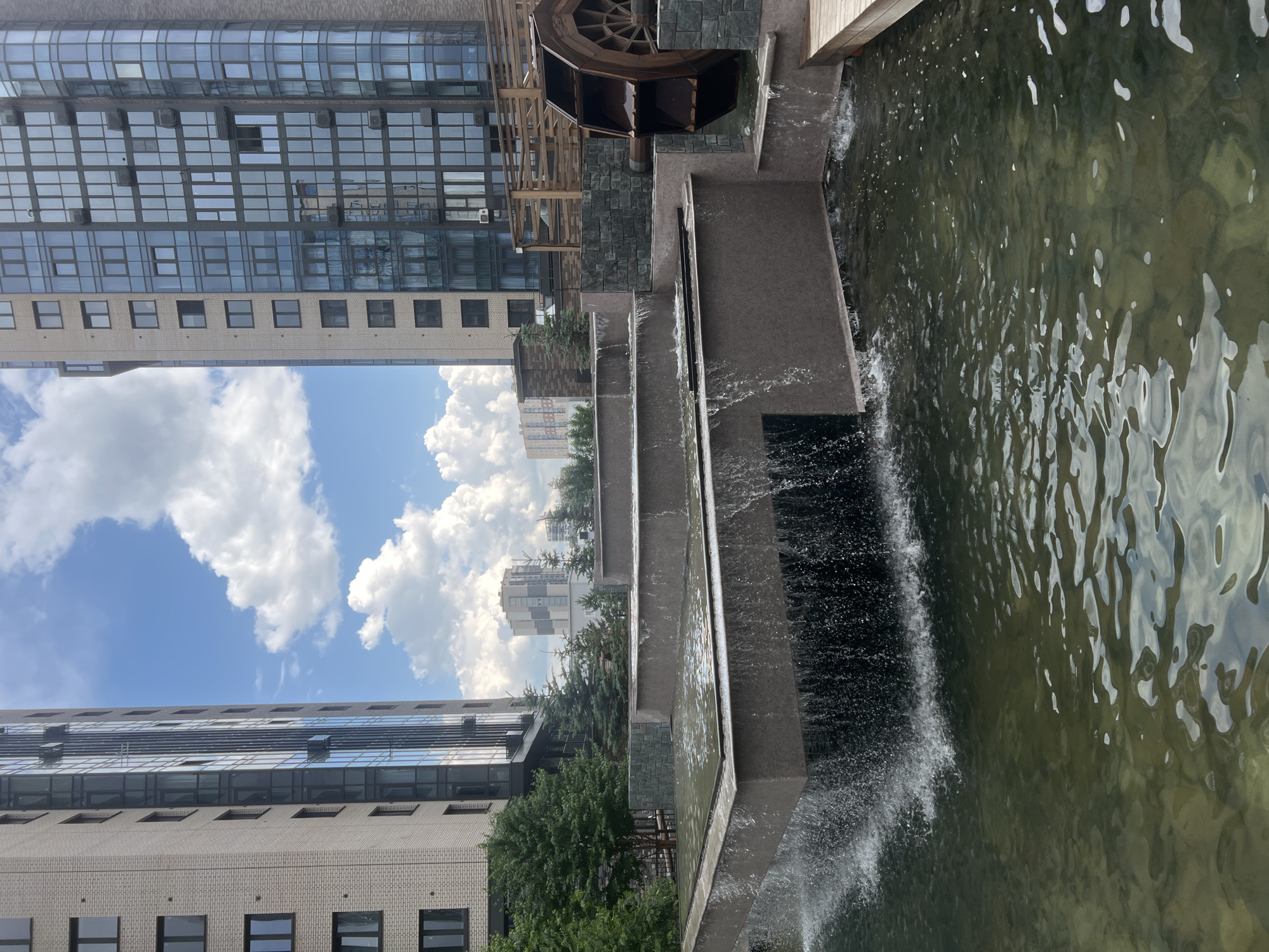 The image depicts a multi-tiered water feature with cascading water in the foreground. The water flows over concrete structures into a pool below. To the right of the water feature, a wooden water wheel is visible, partially submerged. In the background, two modern high-rise buildings dominate the scene, one on the left and one on the right, with numerous windows reflecting the sky and clouds. The sky is blue with scattered white clouds. Green trees and shrubs are present around the water feature and between the buildings. The scene appears to be an outdoor public space, possibly a courtyard or park area within a residential complex. The architectural style of the buildings suggests a contemporary urban environment. The location is identified as Krasnoyarsk, Russia. There are no people visible in the image.