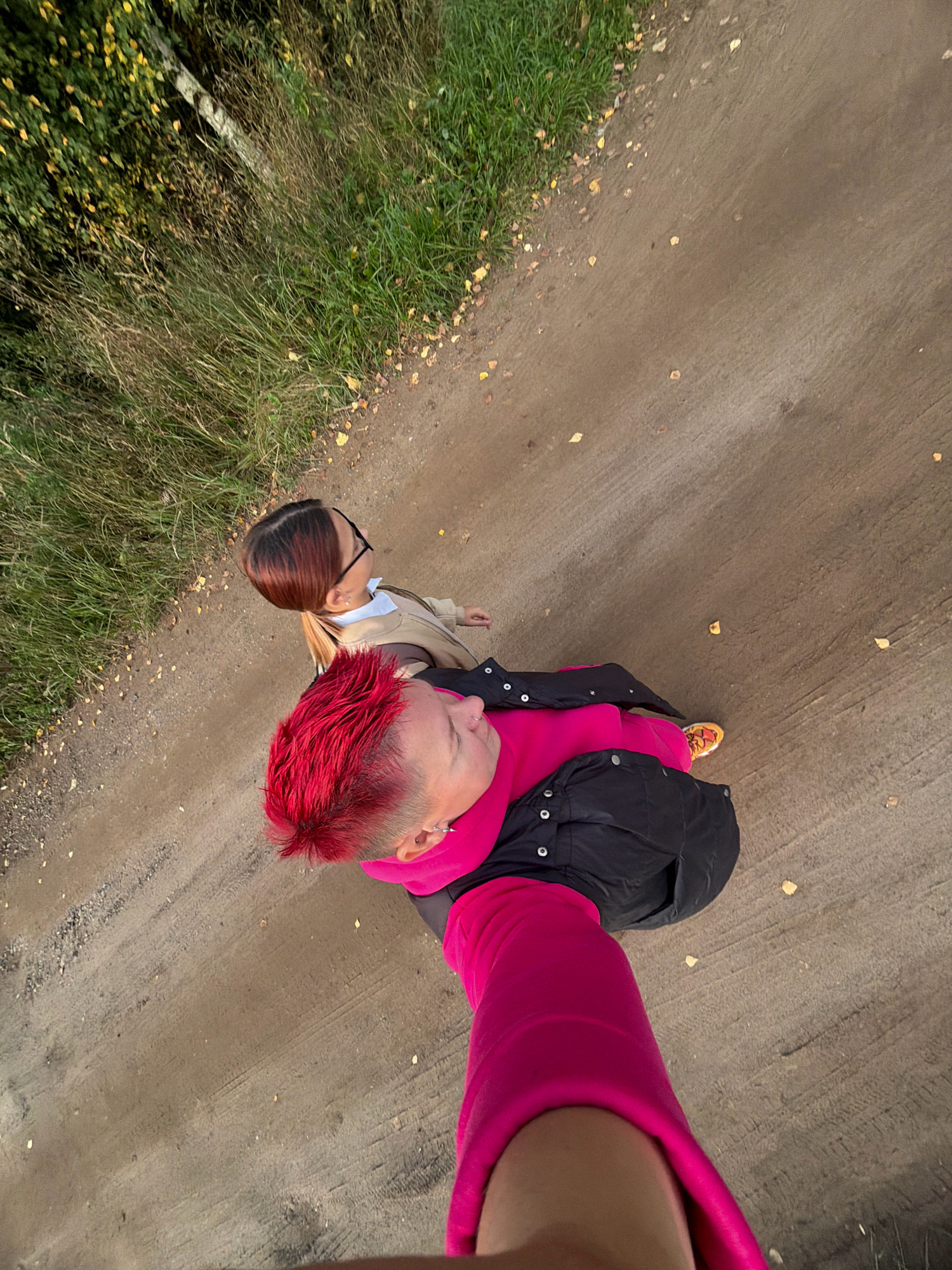 Two individuals are visible from a high-angle perspective, standing on a dirt path. The person in the foreground has bright red, spiky hair and is wearing a black puffer jacket over a bright pink hooded garment. They are also wearing bright orange sneakers with a patterned design. The person in the background has dark hair with blonde highlights pulled into a ponytail and is wearing glasses, a light brown jacket, and a white shirt. The environment appears to be a rural or park-like setting with tall, dry grass and some scattered yellow leaves on the ground. There is a narrow dirt path with some tire tracks visible. The image was taken outdoors. It is not possible to determine the specific location within the city of Chaikovsky, Russia based solely on the visual information provided.