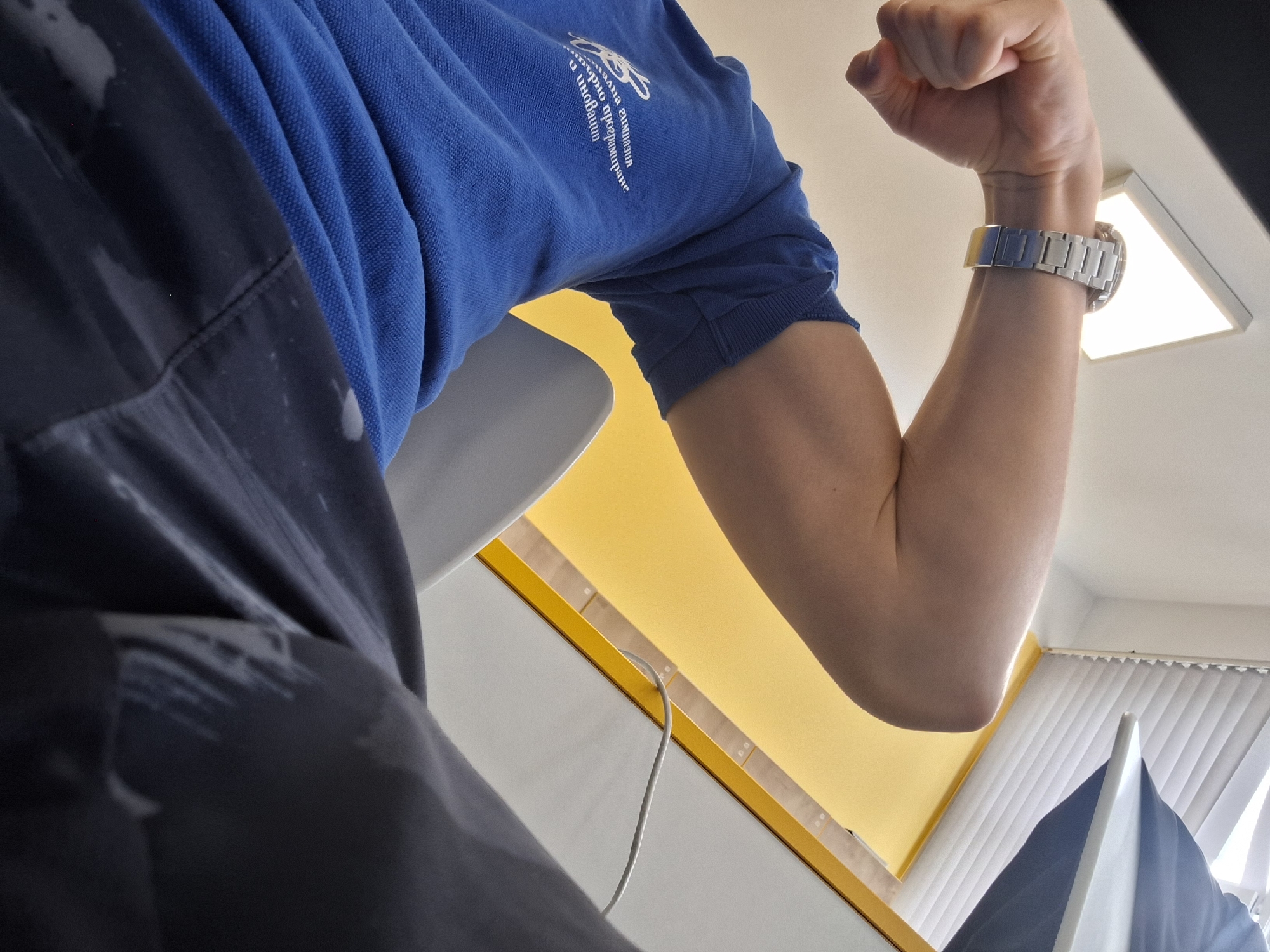A low-angle shot depicts a person flexing their arm. The person is wearing a blue polo shirt with white lettering and design on the left chest. A silver wristwatch adorns their wrist. The person is wearing dark pants. The background includes a yellow and white wall, a white desk with a laptop on it, and white blinds on a window. The scene appears to be indoors, possibly an office or a home office. Burgas, Bulgaria is not evident from the image.