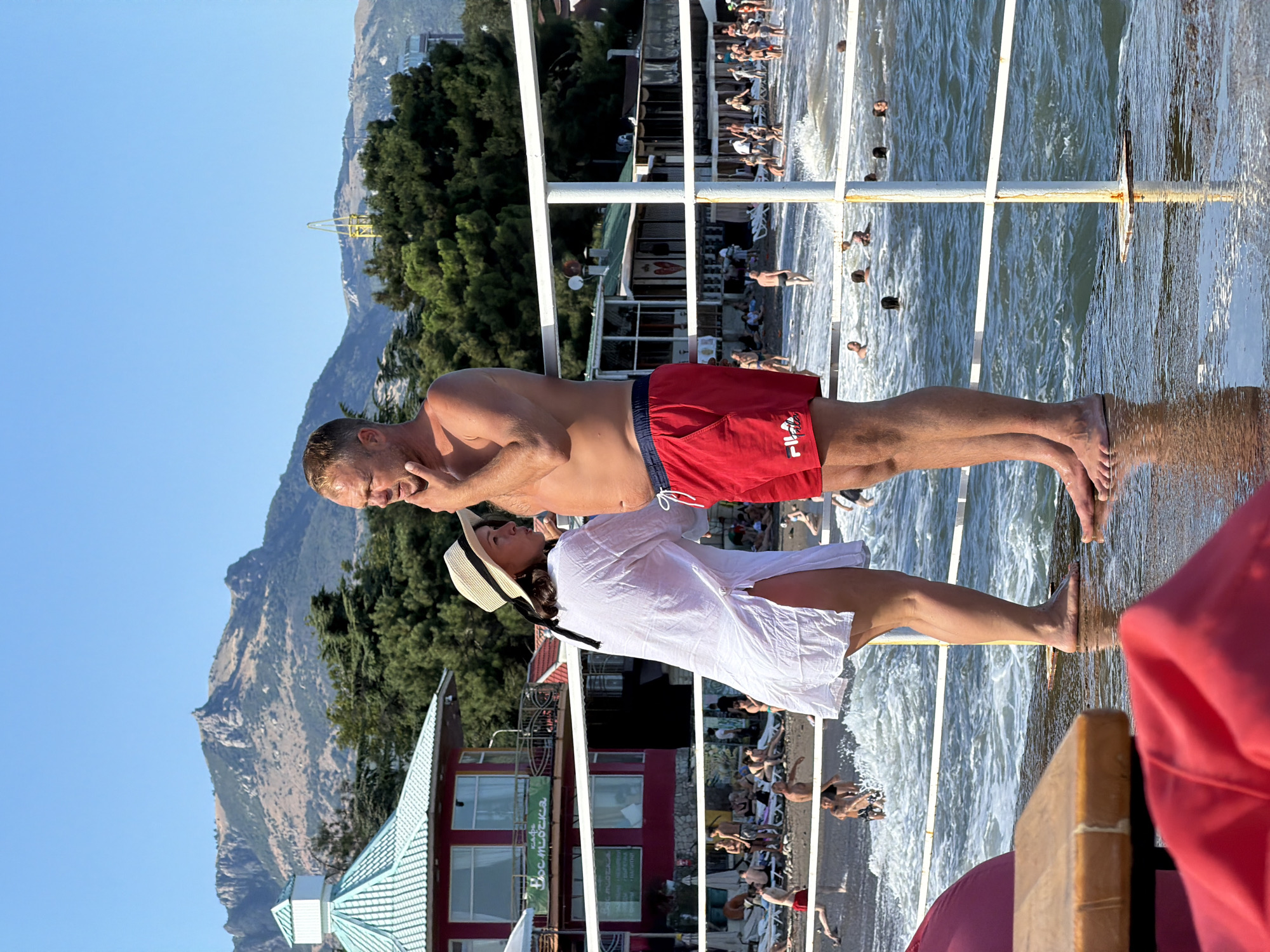 This image depicts a sunny day at a beach in Alushta, Ukraine. In the foreground, a man in red swim trunks and a woman in a white cover-up and straw hat stand on a wet pier, facing away from the camera and looking out towards the sea. The man has his hand on his chin. Behind them, a crowded beach is visible with numerous people sunbathing and swimming in the ocean. Buildings line the beach, and in the background, mountains form a scenic backdrop under a clear blue sky. The water is choppy with waves breaking near the shore.