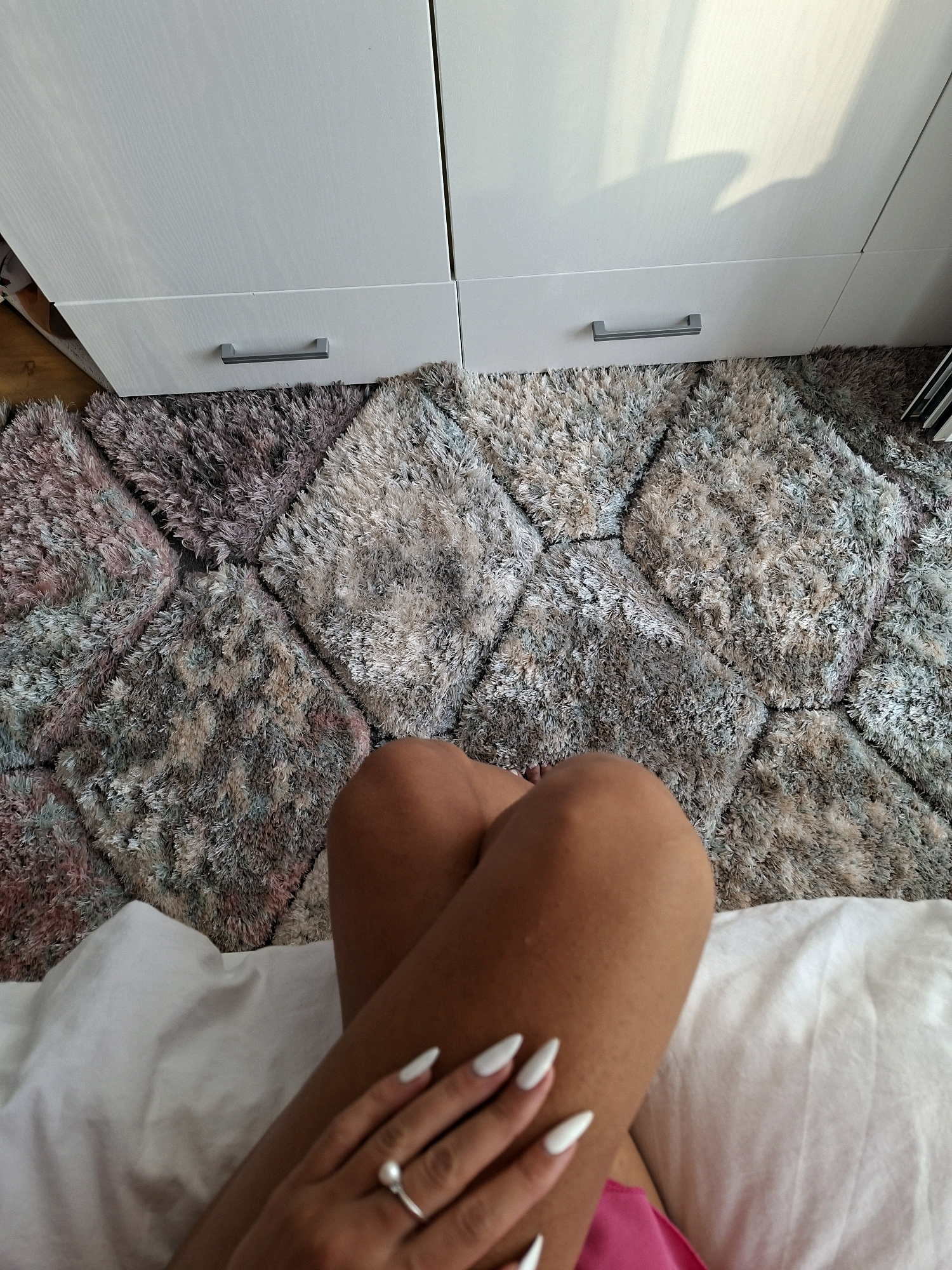 A first-person perspective shot shows a person's legs and hands resting on a bed. The person is wearing a pink garment and a ring with a pearl on their left hand. The bed has white linens and is partially covered by a shag rug with a diamond pattern. The rug is placed in front of a white wardrobe with two doors and two drawers, each with a silver handle. The environment appears to be a bedroom, possibly in an apartment or house.