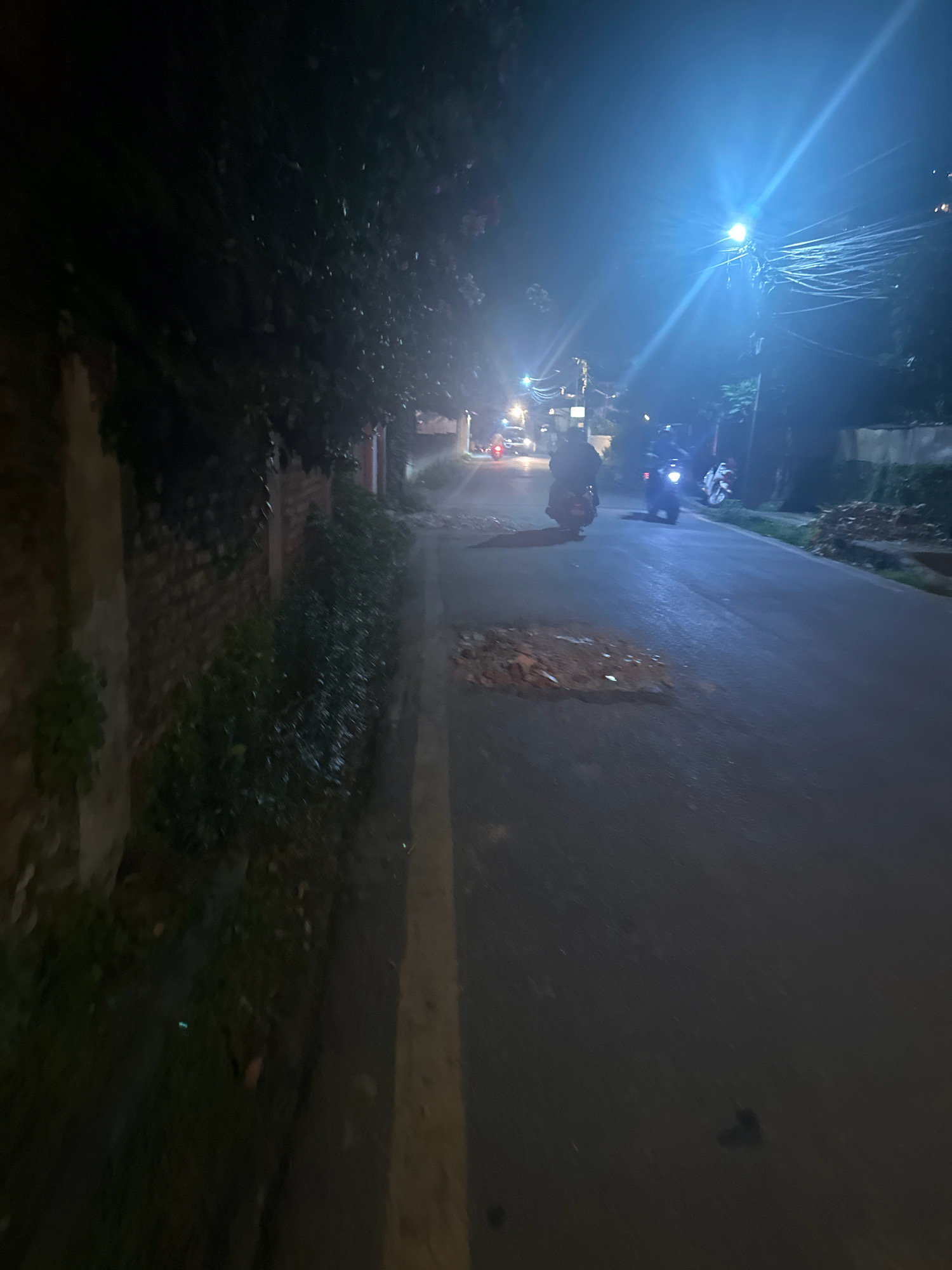 An image taken at night on a paved road in Lalitpur, Nepal, depicts a scene with several motorcycles. The road has a yellow line marking and a significant pothole filled with debris. On the left, a brick wall covered with foliage runs alongside the road. Trees are visible on the left and right sides of the road. A bright streetlamp illuminates the scene from above, casting a strong glare. Two motorcycles are clearly visible on the road, one moving towards the viewer and another away. Further down the road, other vehicles and streetlights can be seen, suggesting a populated area.