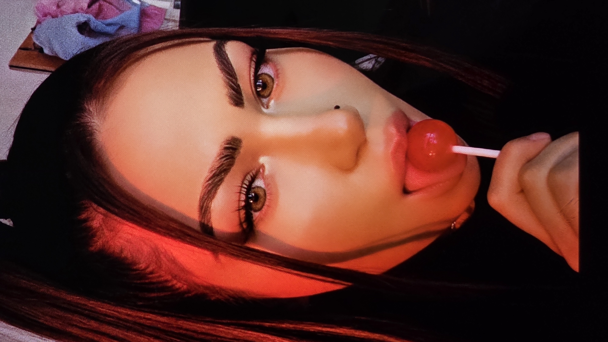 A close-up, angled shot of a woman with brown hair and light skin, holding a red lollipop to her mouth with her right hand. The lollipop touches her tongue, which is slightly extended. Her eyes are brown, and she has a mole on the right side of her nose. The lighting is warm and somewhat saturated, with a red hue on the left side of the frame. In the upper left corner, there are some blurred items, possibly clothing. The background is dark, focusing attention on the woman's face. The location is not discernible from the image.