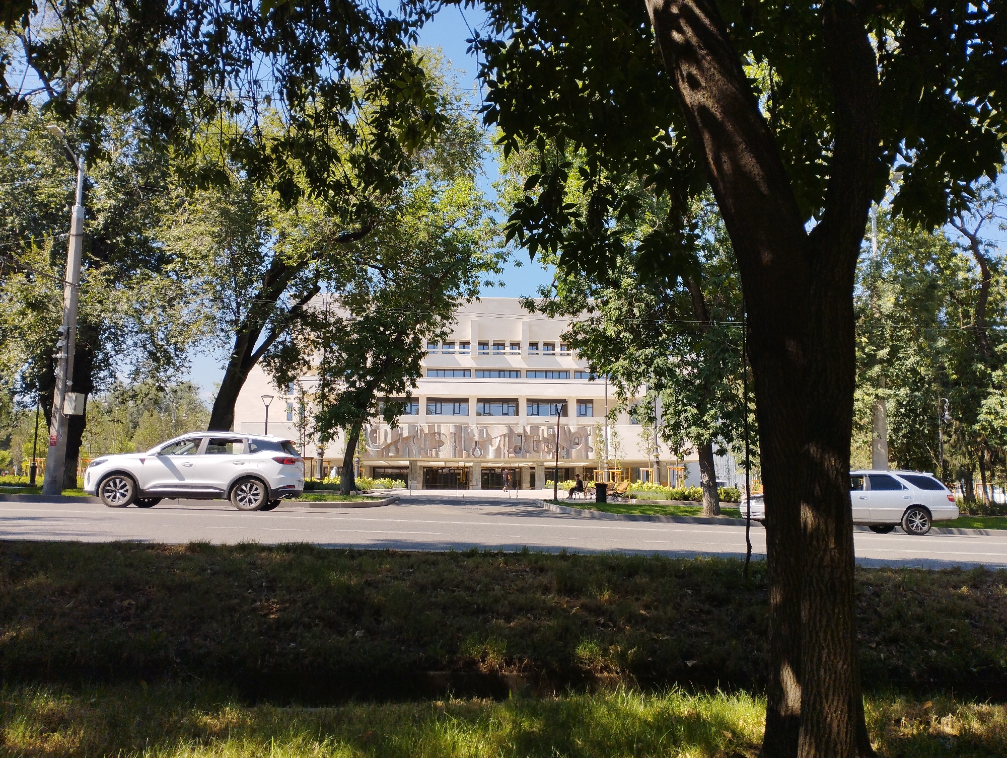 This image depicts a sunny day in Almaty, Kazakhstan, with a view of a large, modern building. The building, likely an educational or cultural institution, features a light-colored facade with numerous windows arranged in horizontal rows across its upper floors. A decorative mural or art installation adorns the lower portion of the building's facade. In front of the building, there is a wide road with two lanes of traffic. A white SUV is driving in the left lane, while a white station wagon is parked or moving slowly in the right lane. The foreground is dominated by a grassy embankment, with several mature trees lining the road and partially obscuring the view of the building and the sky. The trees are lush and green, suggesting it is springtime or summertime. A lamppost is visible on the left side of the road, and there are benches and landscaping in front of the building, suggesting an outdoor public space. The overall atmosphere is serene and urban.