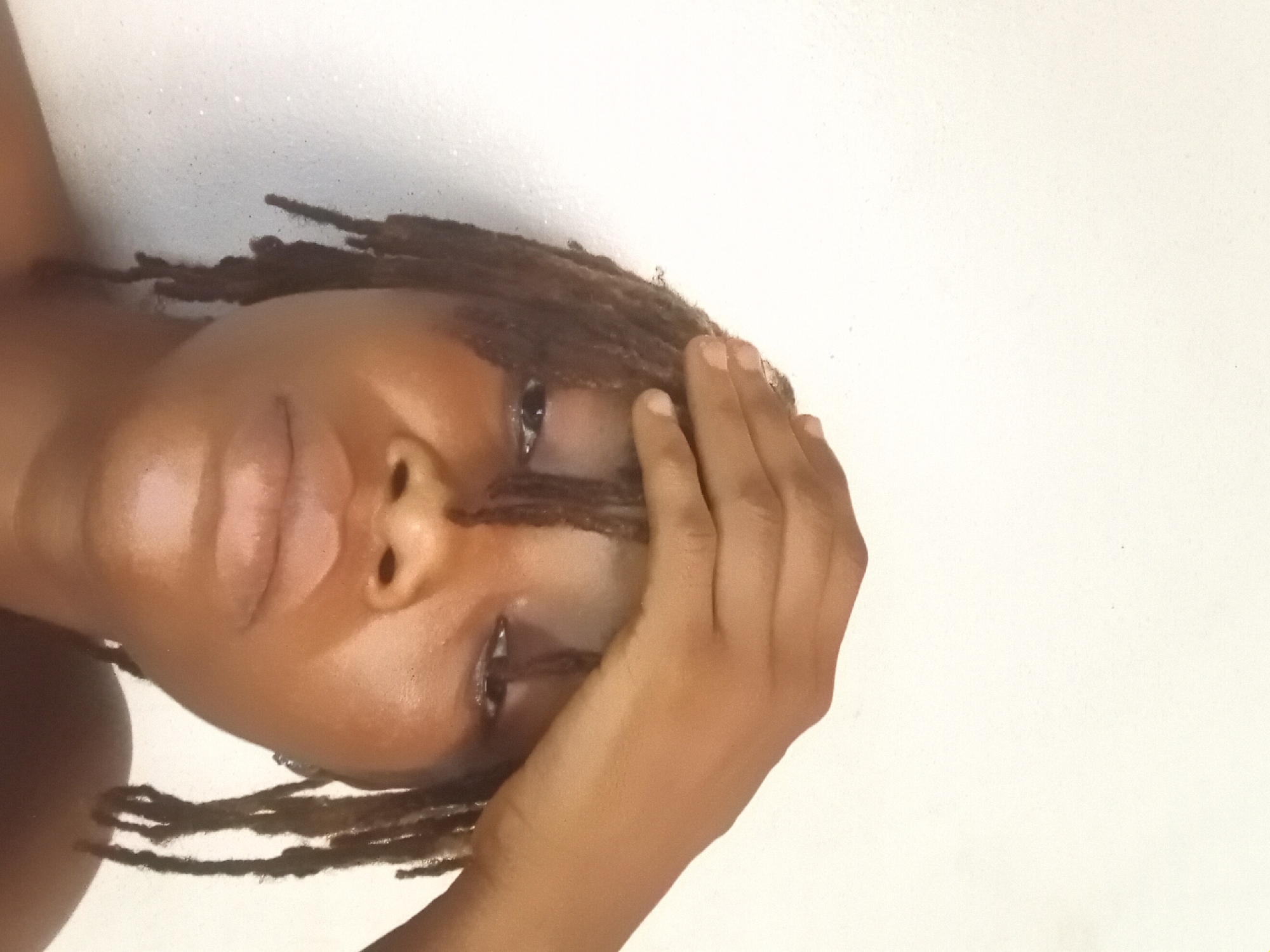 A close-up, angled shot features a dark-skinned person with dreadlocks. The individual is touching their forehead with their right hand. The background is a plain white surface. The location is not discernible from the image; no visual cues indicate it is Shagamu, Nigeria.