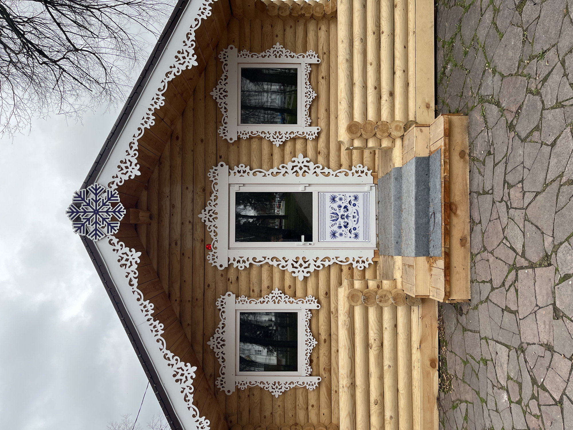 An exterior shot showcases a charming, newly constructed or well-maintained log cabin-style house in Ulyanovsk, Russia. The house is built from light-colored, neatly stacked logs, presenting a rustic yet ornate appearance.
The primary subjects are the architectural details of the house. Two windows and a door on the facade are framed by elaborate, white carved wooden decorative elements, known as nalichniki, which feature intricate scrollwork and floral patterns. A prominent dark blue and white snowflake ornament adorns the peak of the gable, complementing the white trim along the roof's eaves. The main entrance features a white door with a window section, below which is a white panel decorated with blue folk-art patterns, reminiscent of traditional Russian designs like Gzhel. A simple wooden step structure with grey treads leads up to the door.
The setting appears to be a residential or public area, with a textured stone pathway, featuring irregular grey flagstones and patches of green grass, leading up to the house. The sky above is overcast and grey, and the bare branches of a deciduous tree are visible against it, suggesting a late autumn, winter, or early spring season. The reflections in the windows also show the bare trees and cloudy sky. There are no people visible, and no specific activity is taking place other than the static display of the building. No visible text is present in the image.