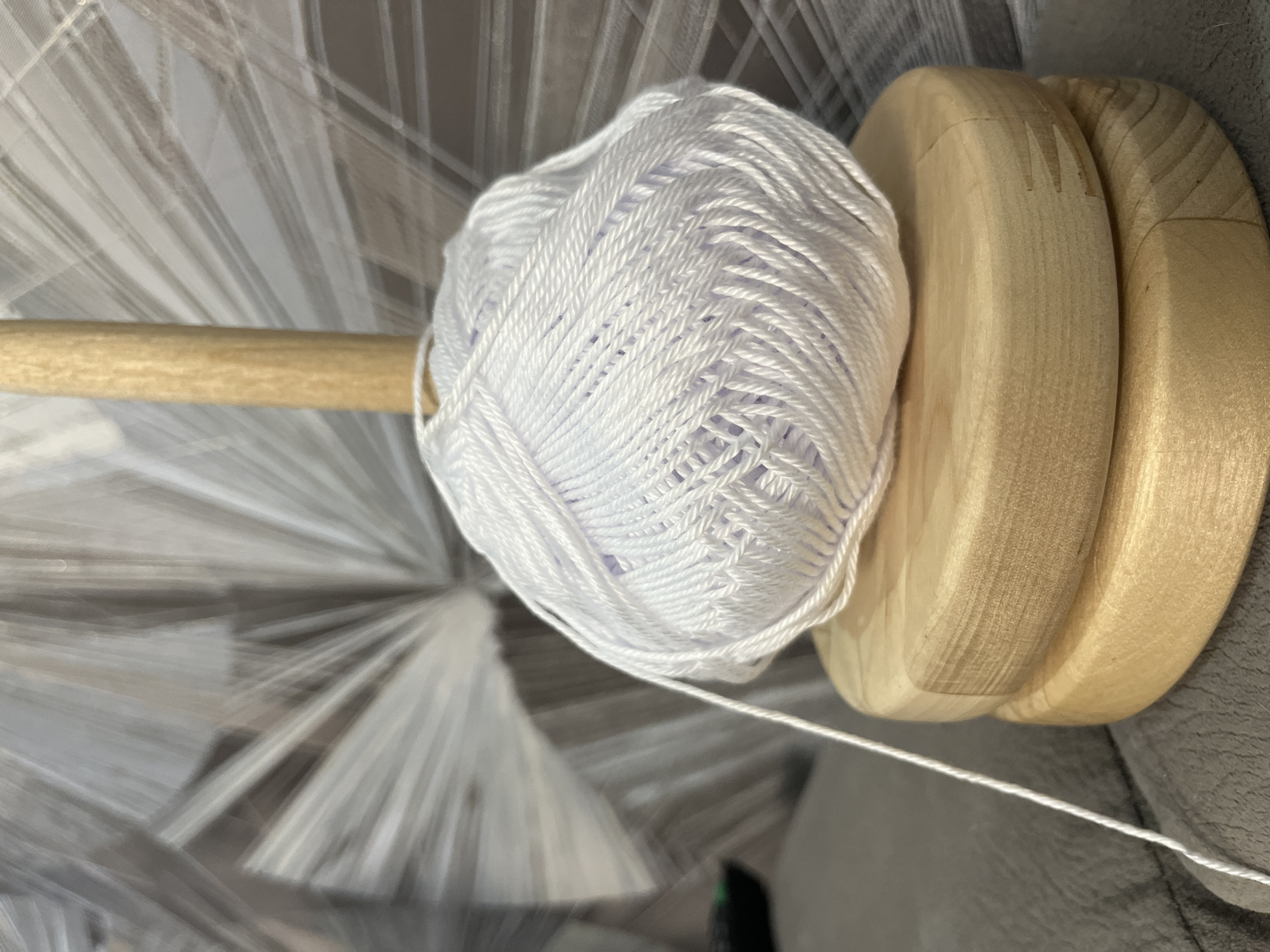 A close-up shot of a ball of white yarn sitting on a wooden yarn holder. The yarn is tightly wound and textured. The wooden yarn holder has three rounded, stacked sections. A thin strand of yarn extends from the ball, disappearing out of frame. The background consists of blurred, white and gray patterns that suggest a soft or draped material. No people are visible. The city of Chaikovsky, Russia is not discernible in this image.