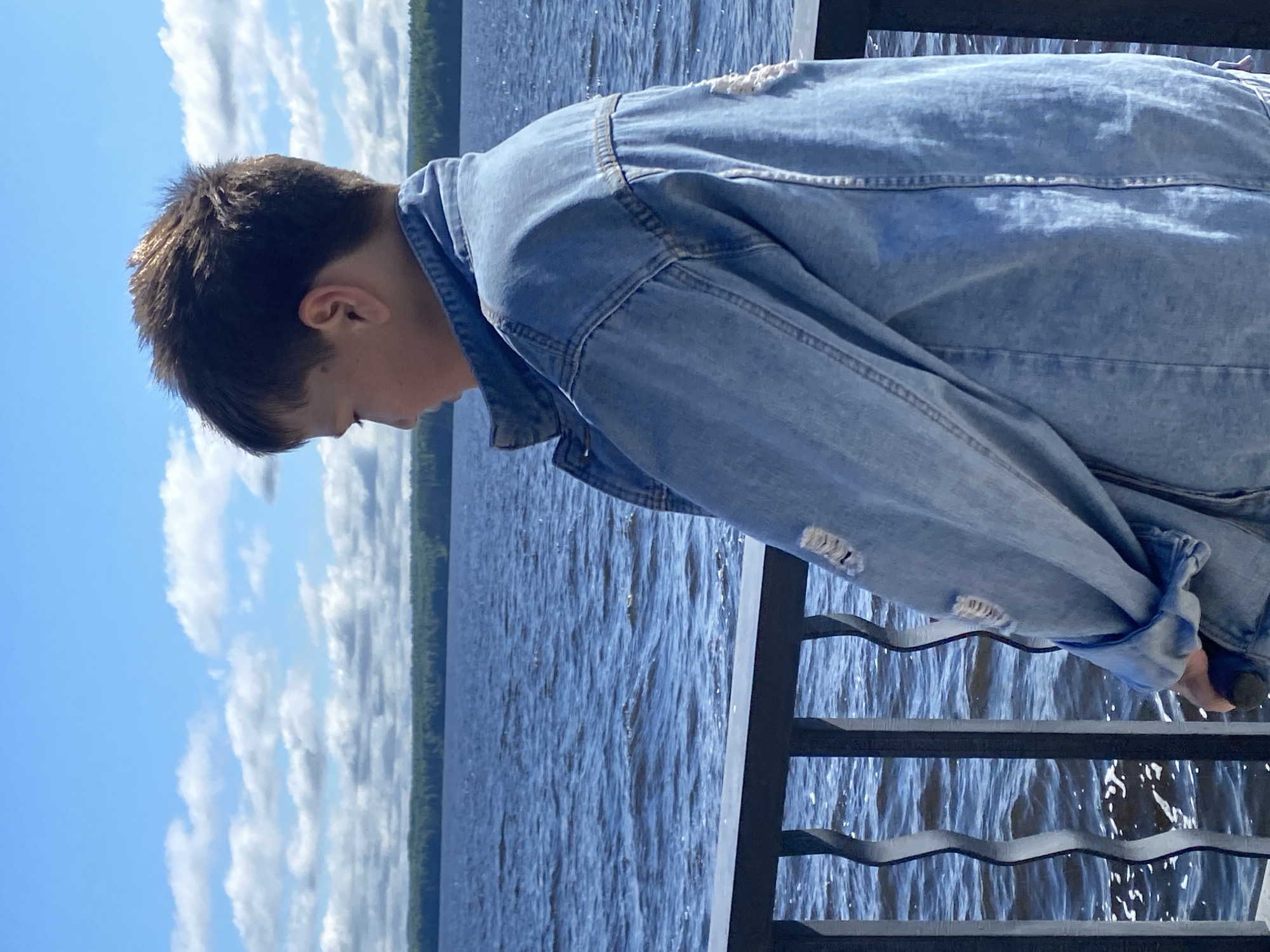 A young boy, viewed from behind and to the side, stands on a balcony overlooking a large body of water. He is wearing a light blue distressed denim jacket. The water is choppy, with whitecaps indicating wind. In the distance, a tree line marks the opposite shore. The sky above is blue with scattered white clouds. The balcony railing is black and has a decorative, wavy design. The scene appears to be set on a sunny day, possibly in Chaikovsky, Russia, given the presence of a large reservoir that is characteristic of the area.