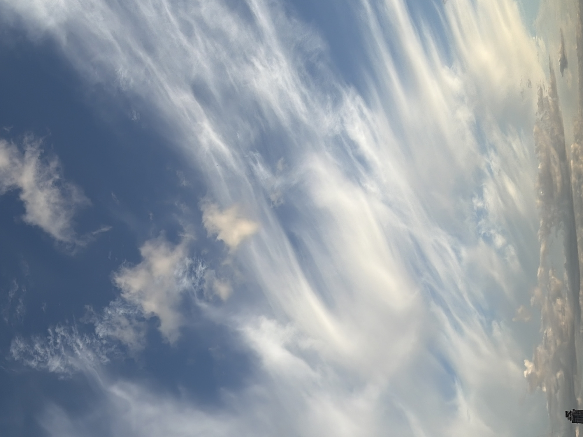 The image captures a vast expanse of blue sky filled with wispy white clouds. The clouds are stretched and layered, creating a sense of movement and depth. Some of the clouds are thin and feathery, while others are more substantial and textured. The light filtering through the clouds suggests a daytime scene. At the very bottom left of the image, a portion of a tall, dark building is visible, suggesting an urban environment, possibly Khimki, Russia, given the context provided. The rest of the image is dominated by the sky and clouds, with no other discernable objects or people.